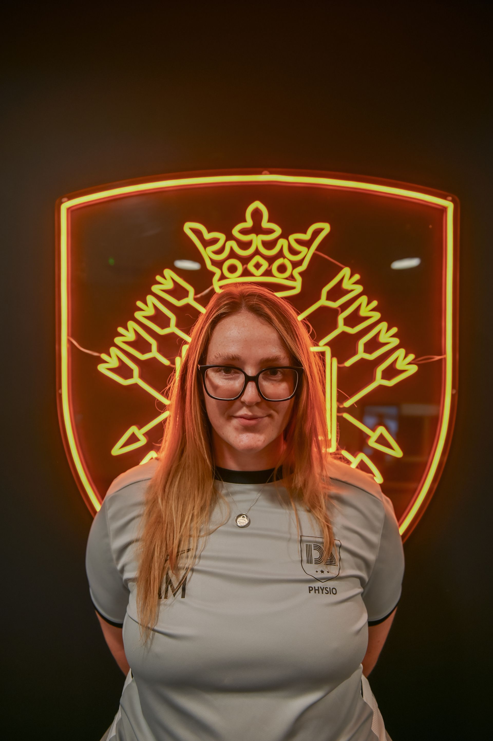 Woman in glasses stands before a glowing yellow shield logo on a black background.