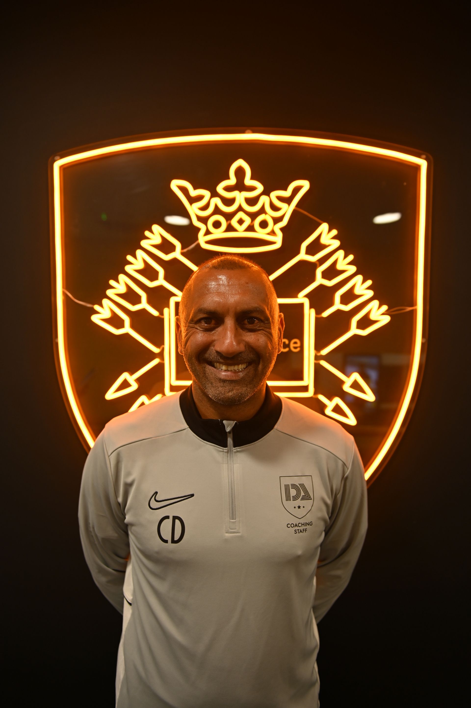 Man smiling, wearing a gray shirt, stands in front of a neon-lit crest.