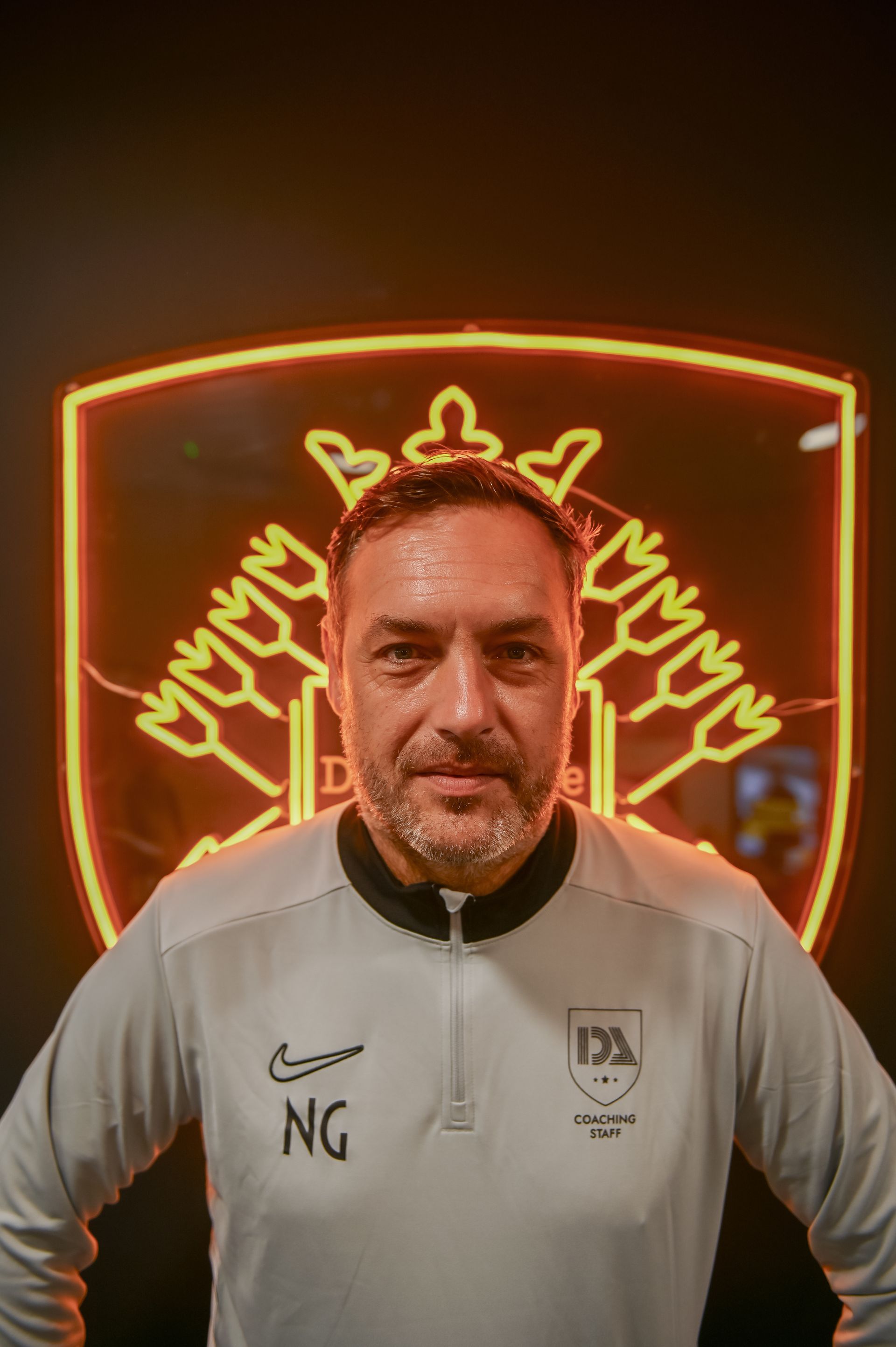Man in light gray jacket, standing in front of illuminated team emblem.