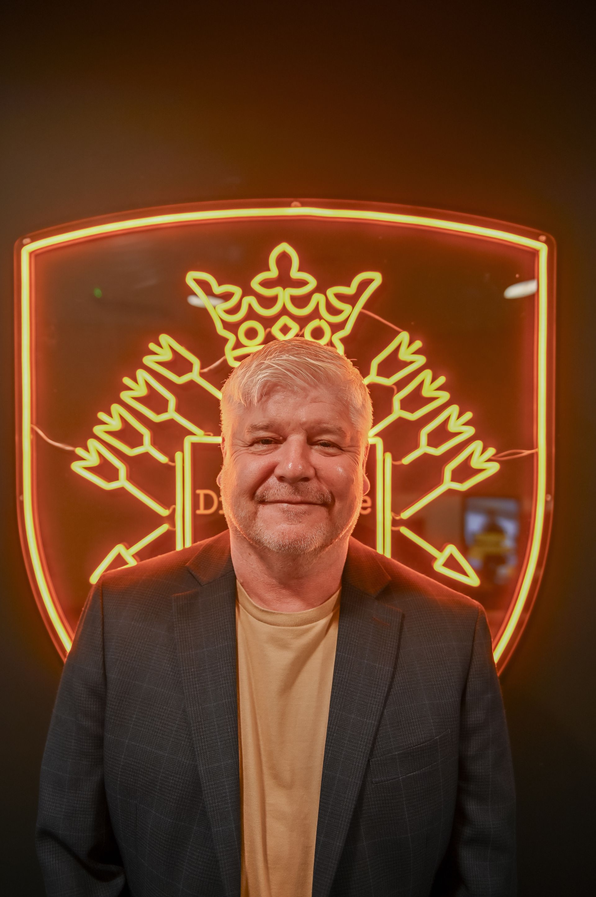 Man in blazer stands before a neon crest with crown, arrows, and 