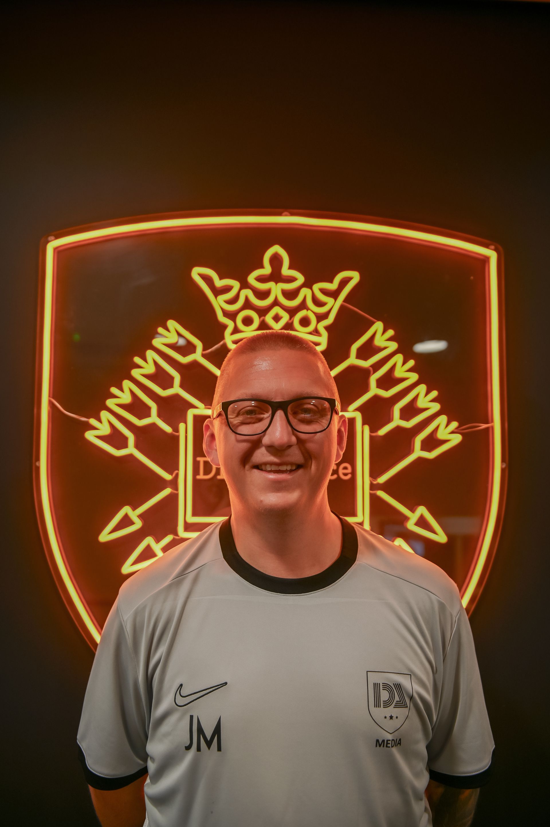 Man in glasses smiling, in front of a neon emblem.