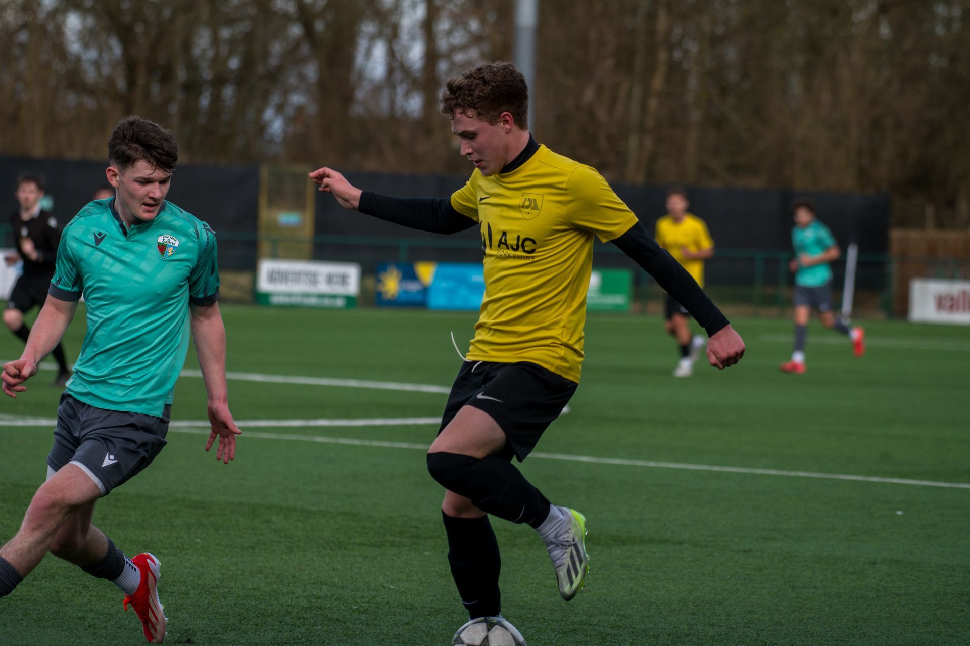 Soccer players on a field. A player in a yellow jersey dribbles the ball while another in a green jersey defends.