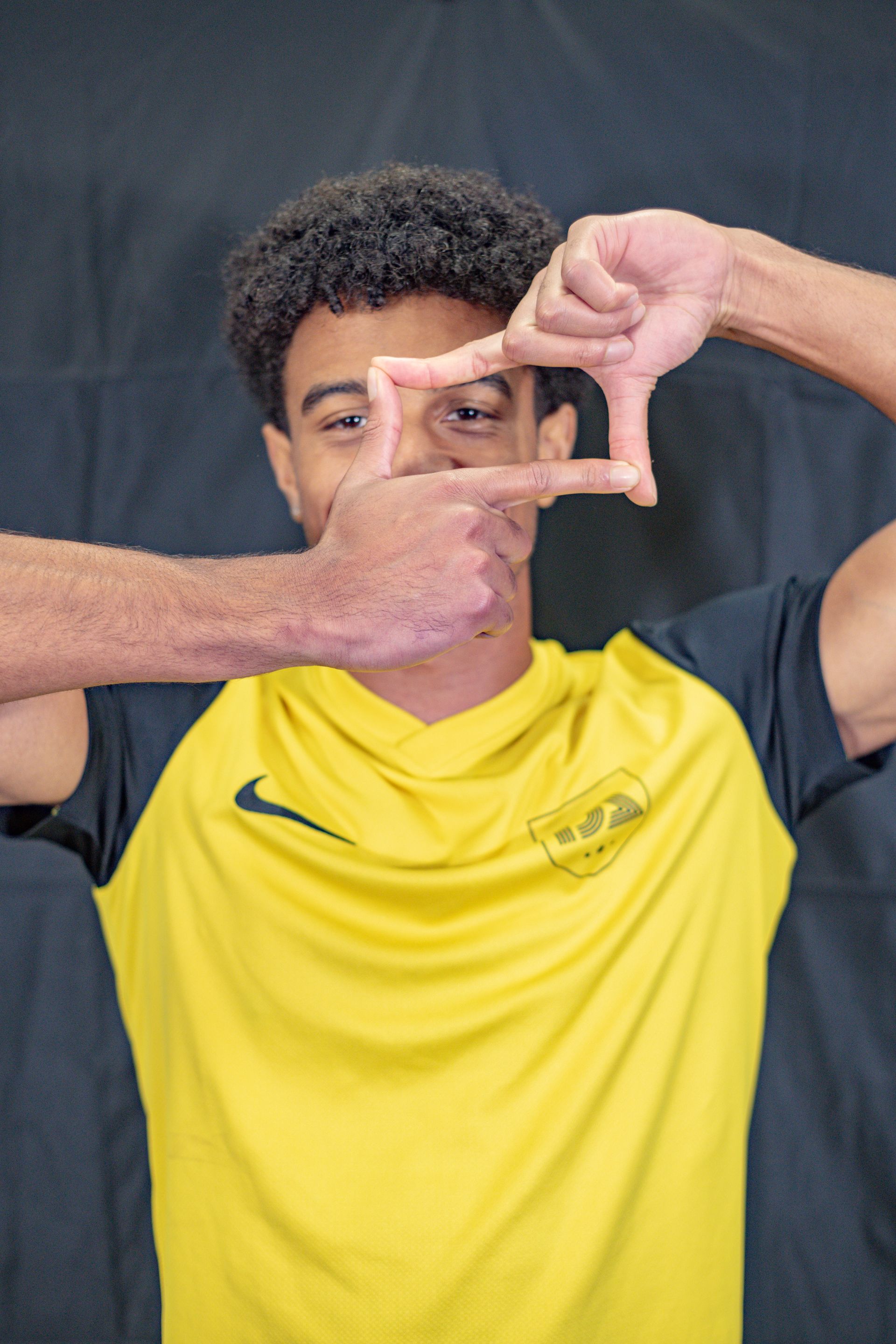 Man in yellow and black shirt making a frame with his hands, smiling, in front of a dark backdrop.