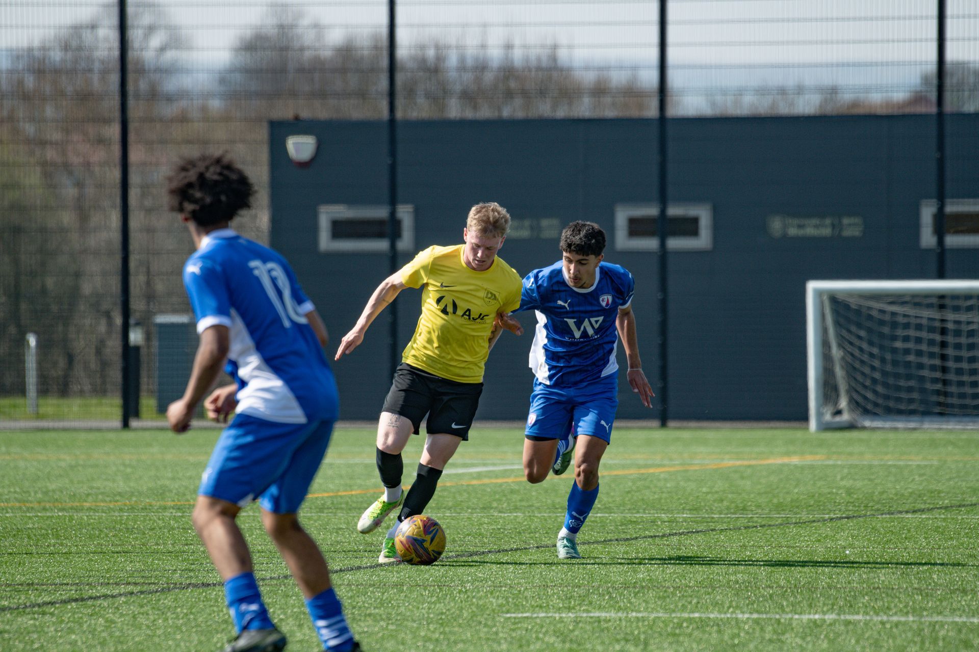 Soccer players in a game on a green field. A player in yellow dribbles the ball, pursued by blue-clad opponents.