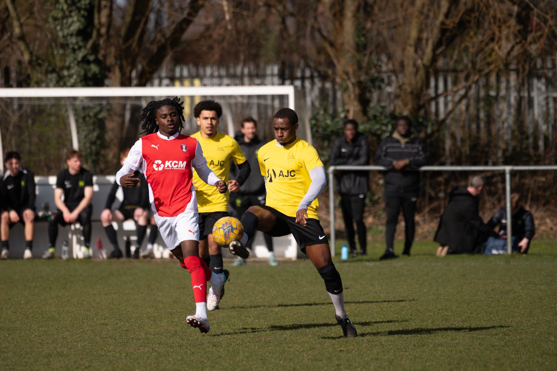 Soccer players in red and yellow jerseys on a sunny field, one dribbling a ball.