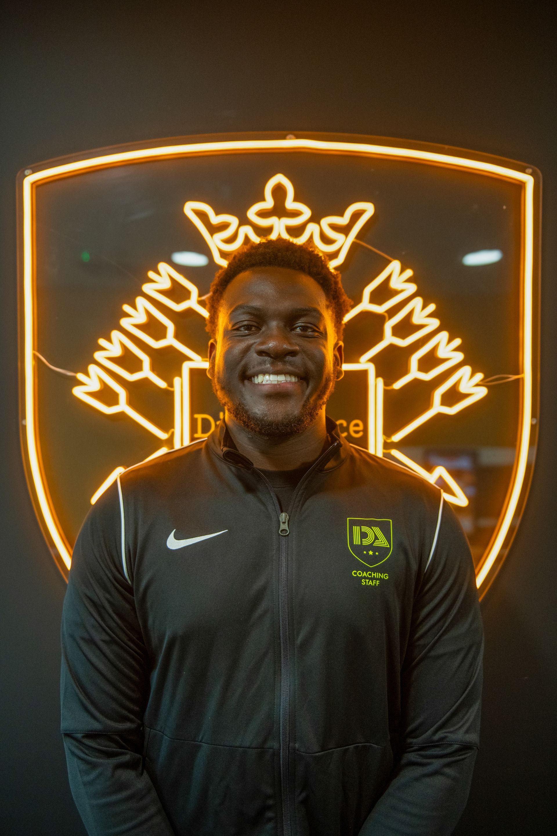 Man smiling in front of a neon logo, wearing a black jacket with a logo.