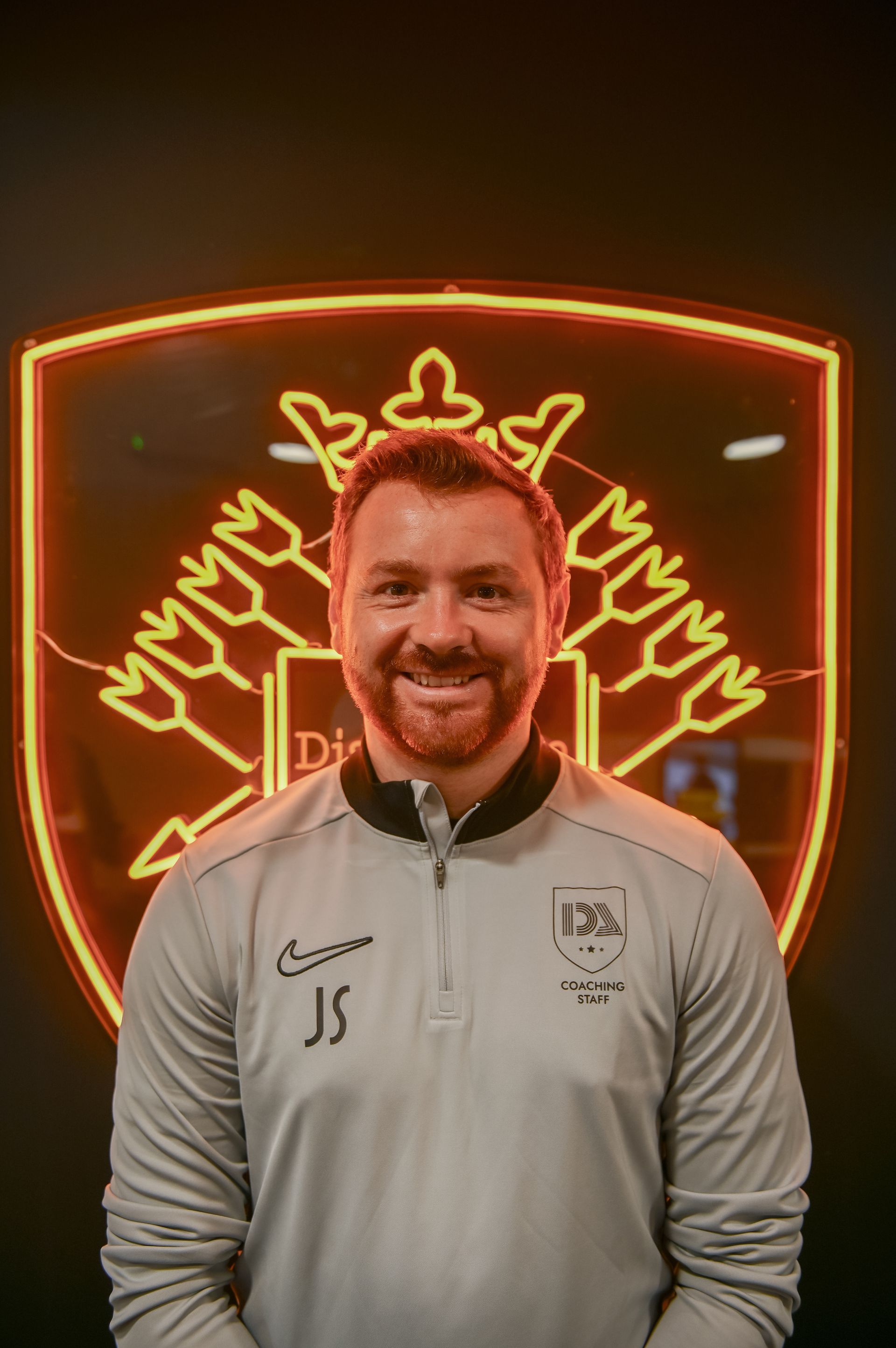Man with beard smiles in front of a neon logo. He wears a grey jacket with 