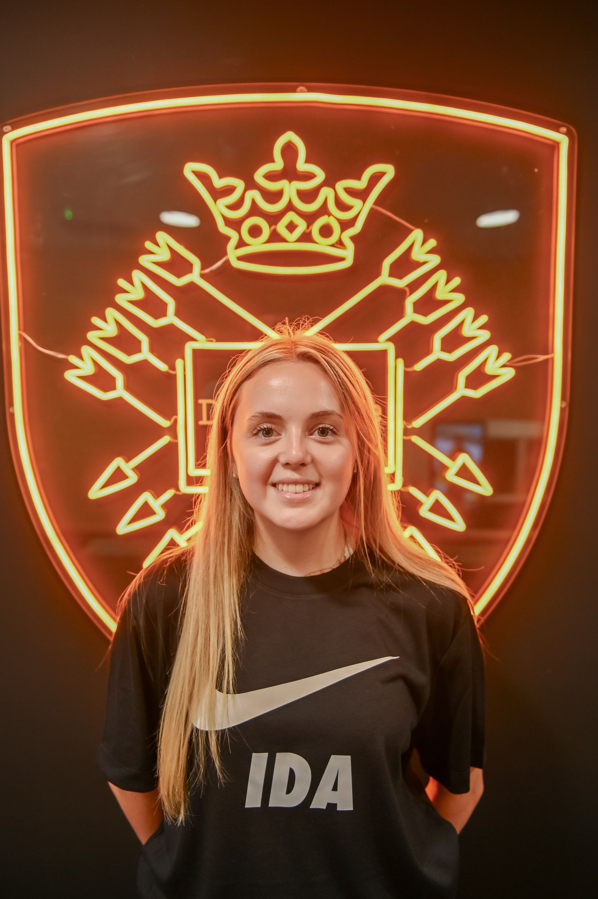 Woman in black shirt with logo smiling in front of a neon emblem.