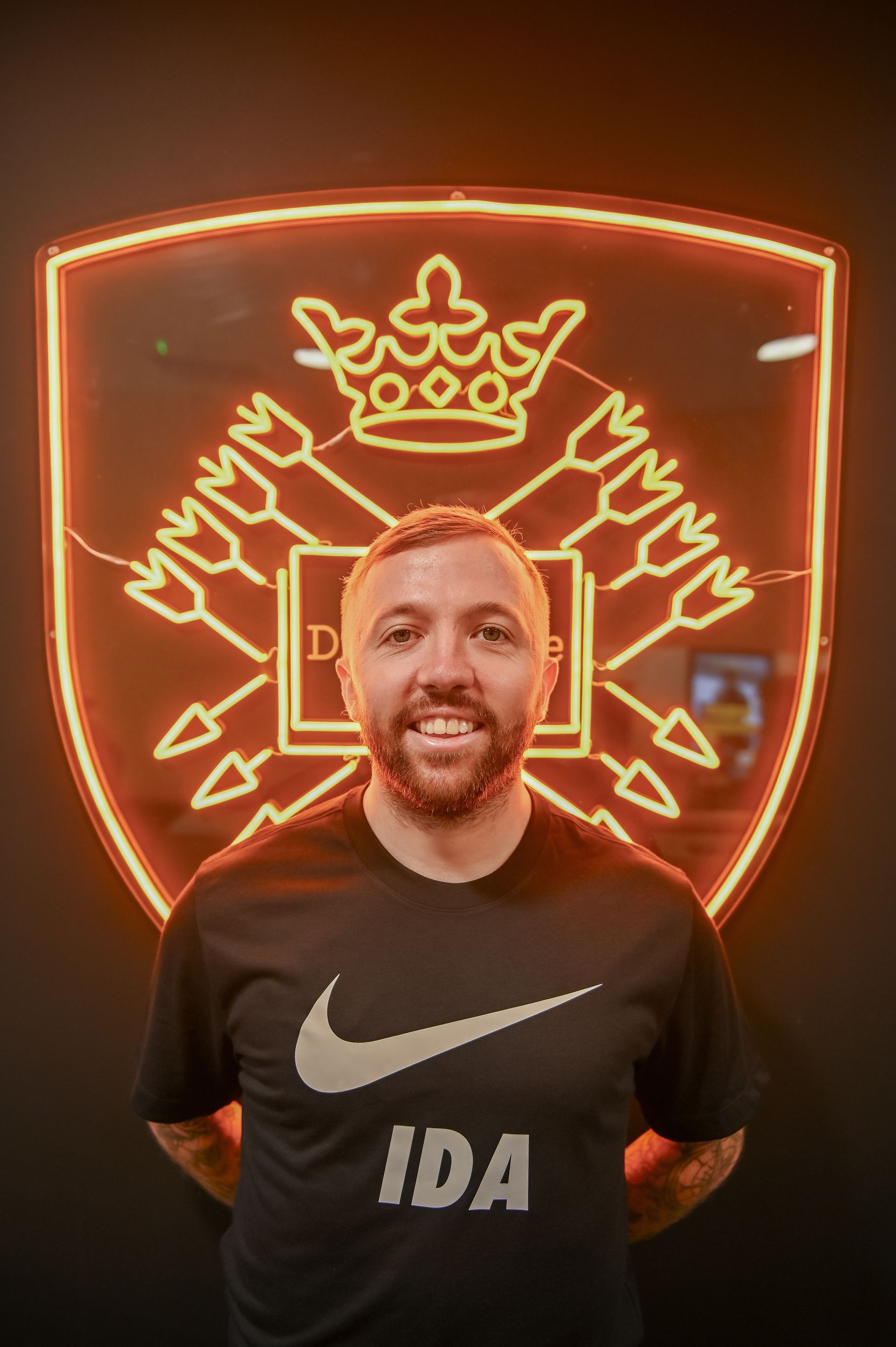 Man in black t-shirt smiles in front of a neon logo, which is a crown with arrows.