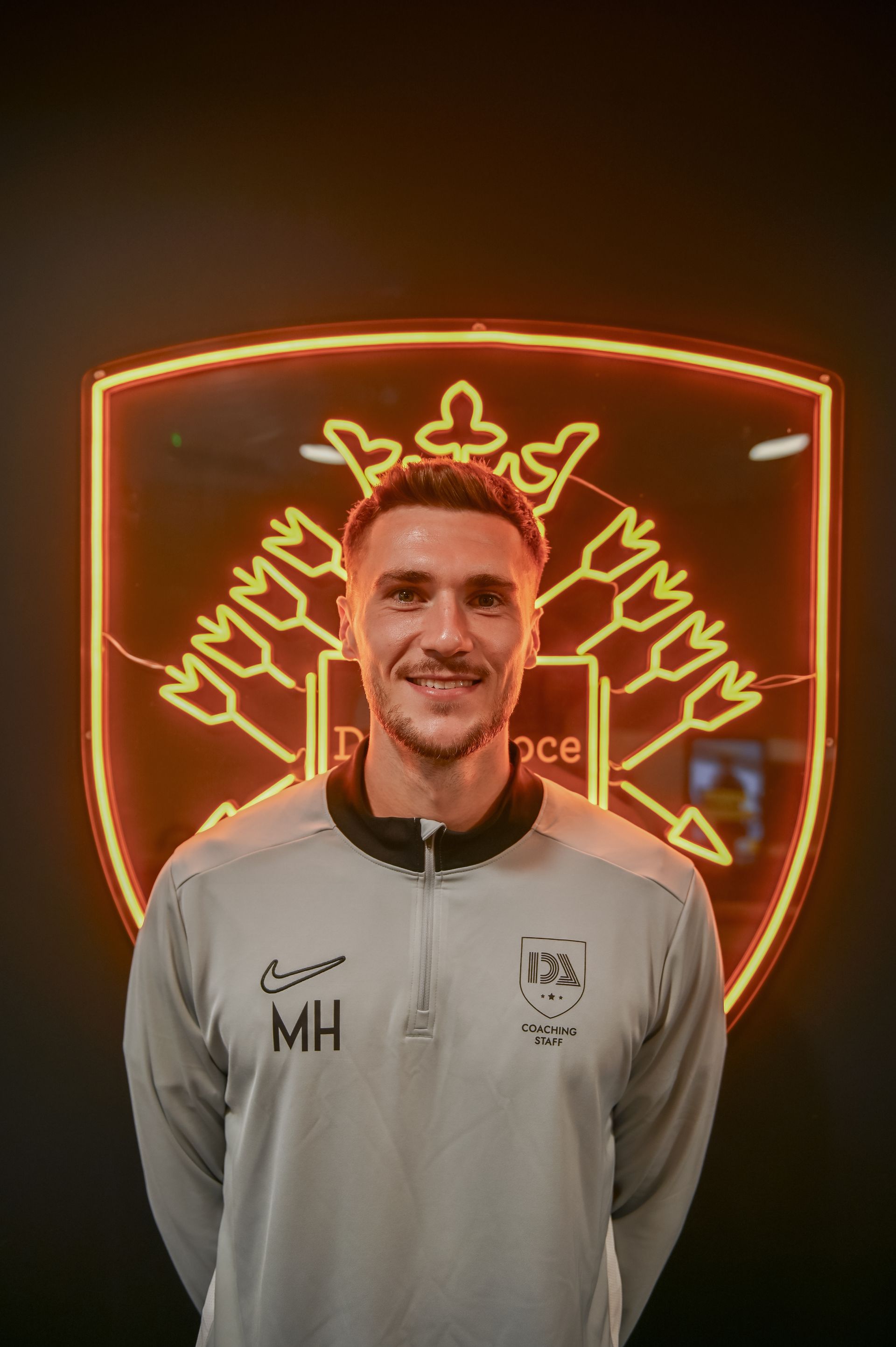 Man in a light gray athletic jacket smiles in front of a neon illuminated crest.