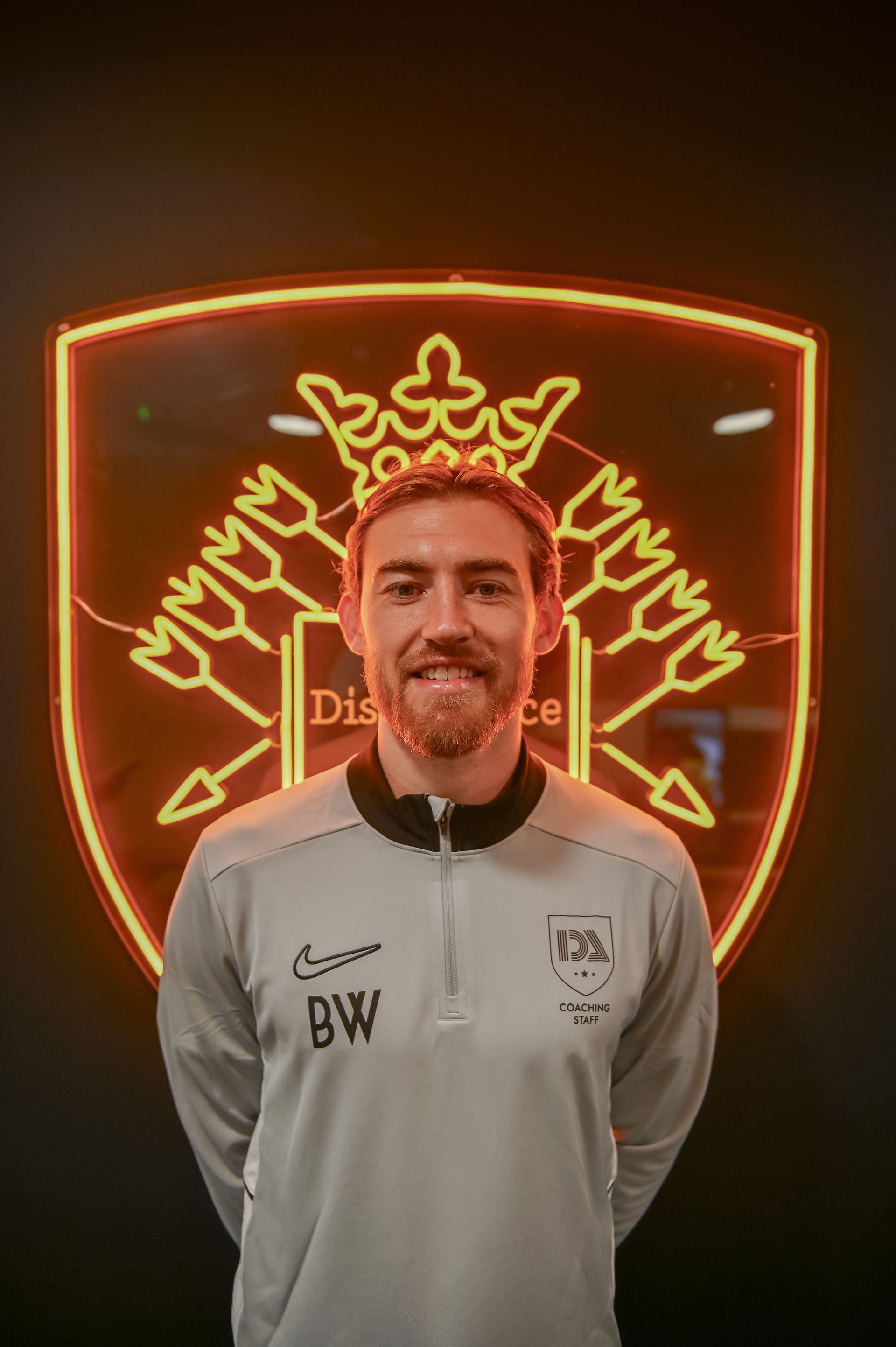 Man in grey sportswear stands in front of a neon illuminated crest. He smiles, with 