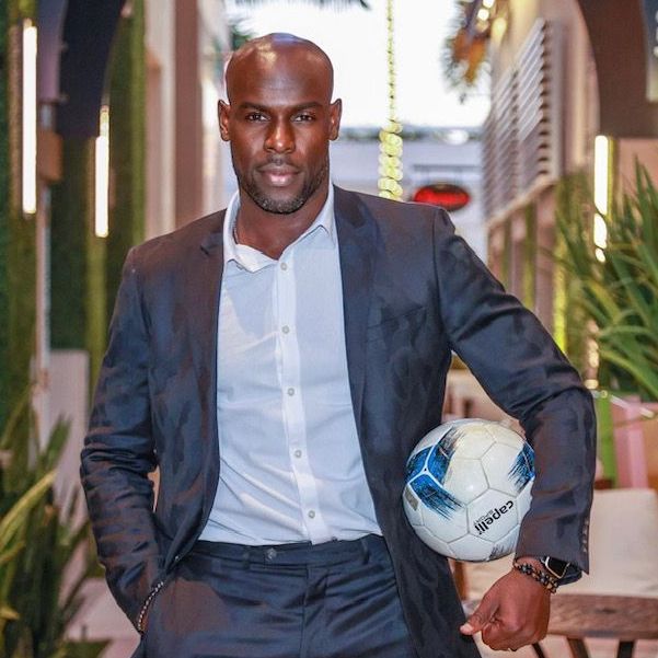Man in a suit holds a soccer ball, standing in an outdoor walkway with greenery.