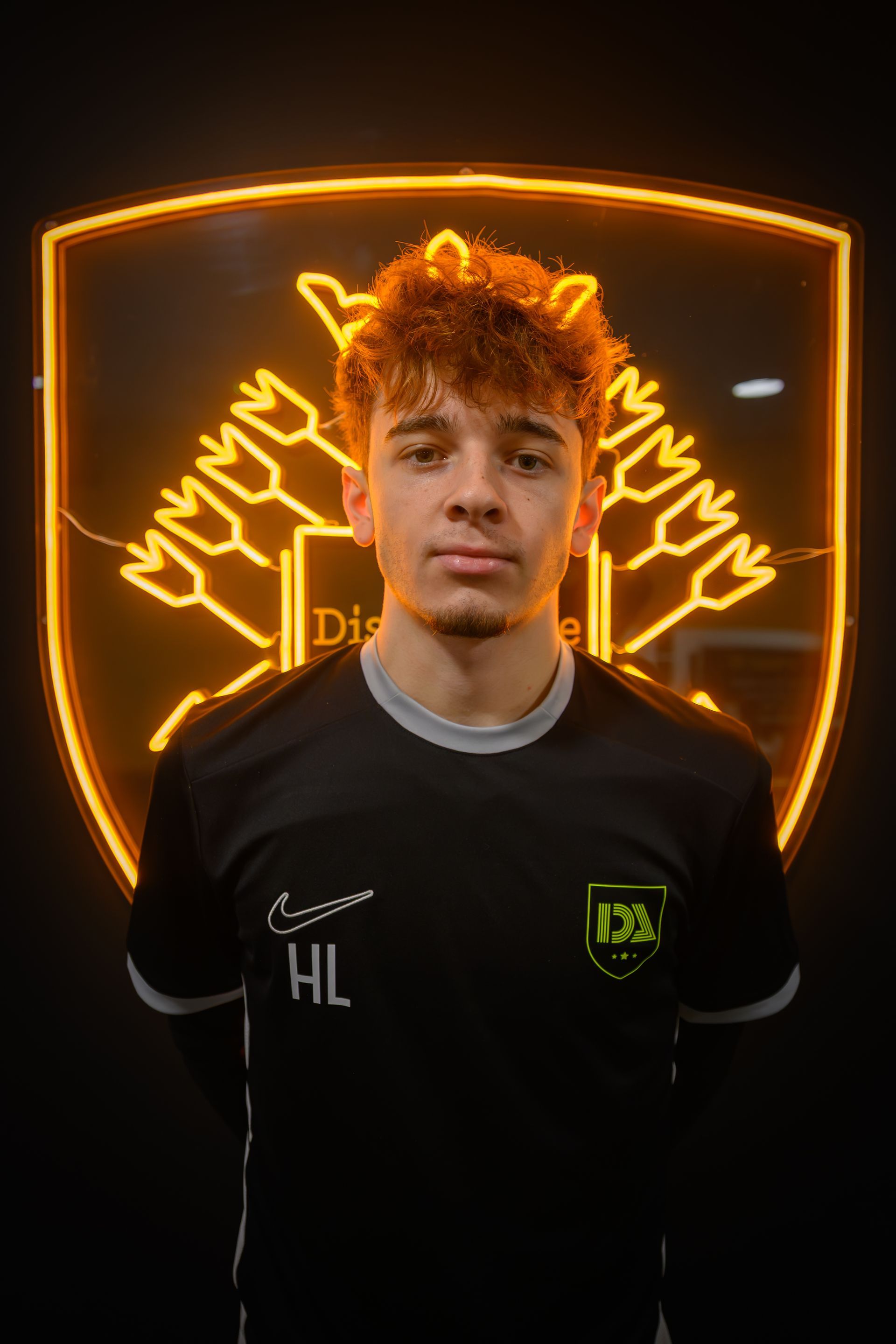 Young man in black shirt stands in front of a neon logo, arms at sides.