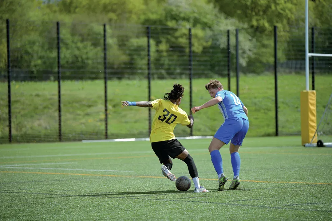 Two soccer players on a green field: one in yellow, kicking the ball; the other in blue, reaching.