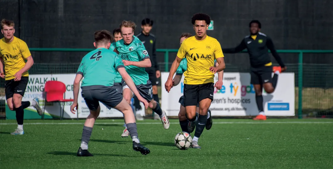 Soccer players in yellow and teal uniforms play on a green field. A player dribbles the ball toward a defender.