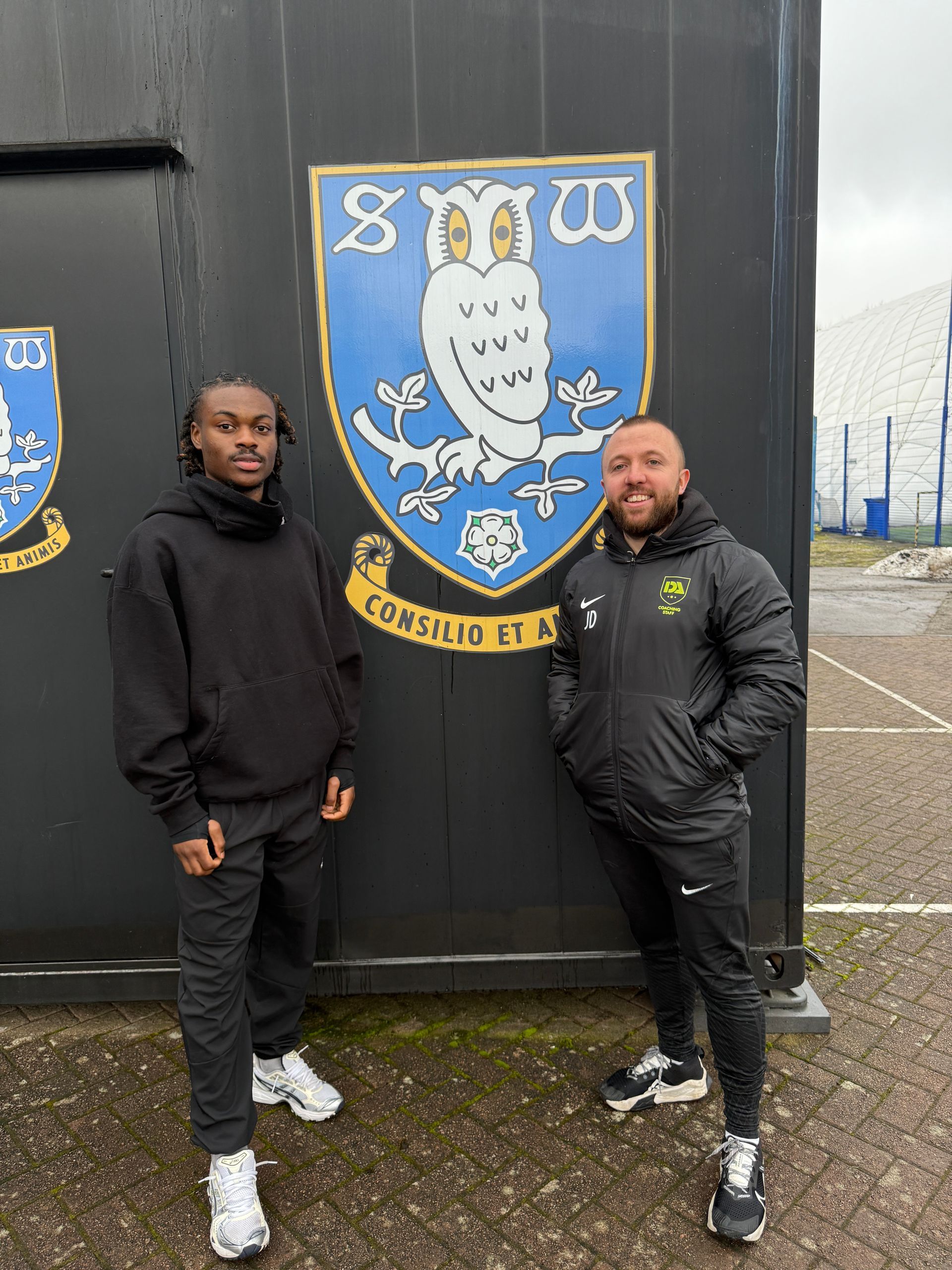 Two men stand in front of a blue and white emblem with an owl, black building background.