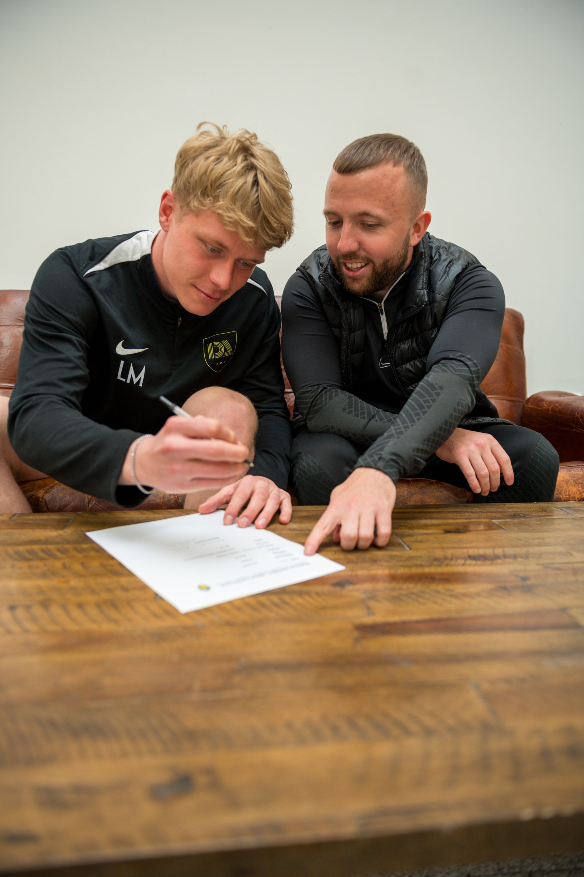 Two men at a table, one signing a document as the other points.