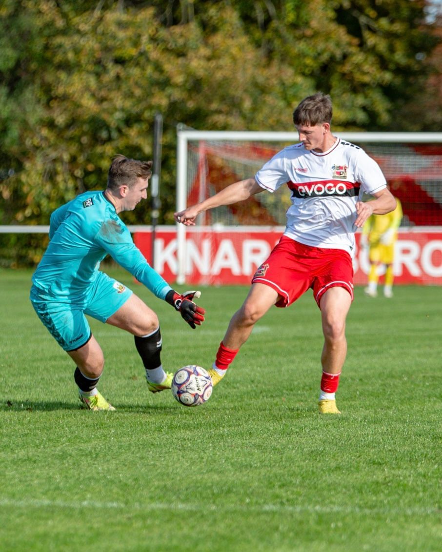 Soccer player kicking ball, goalkeeper dives, both in bright uniforms on green field.