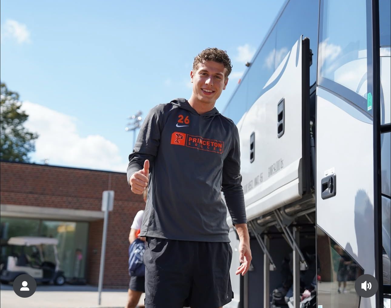 Man in orange sweatshirt giving a thumbs-up next to a bus, smiling.