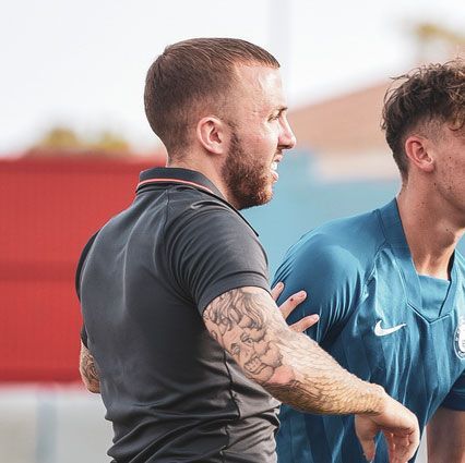 Man with arm tattoos and polo shirt, talking to a person in a blue shirt on a field.