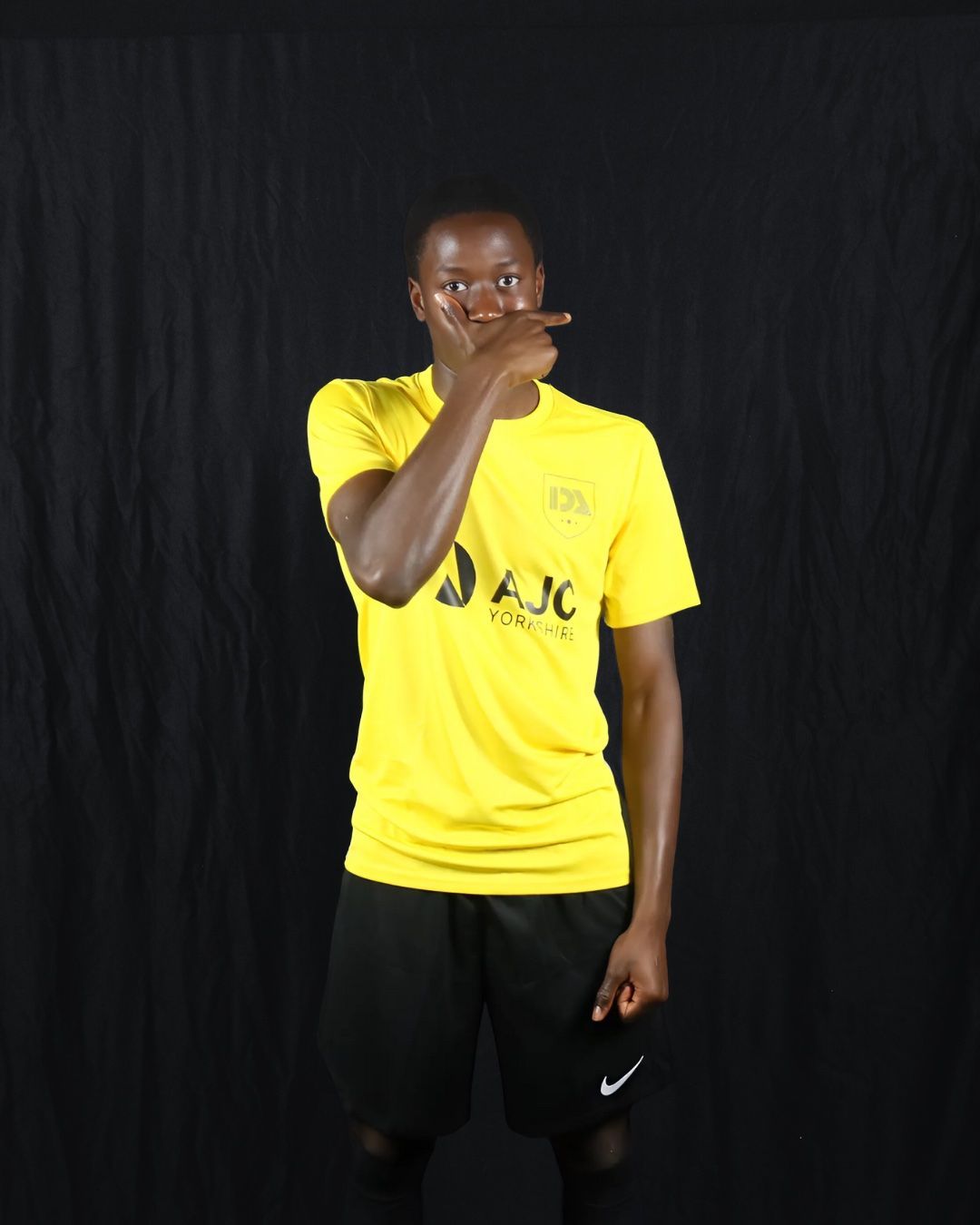 Man in yellow shirt and black shorts points at his temple with a black backdrop.