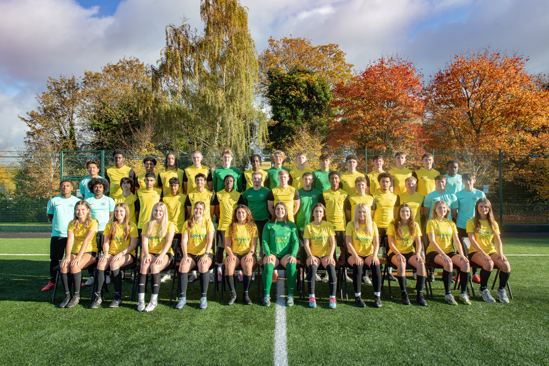 Soccer team in yellow uniforms poses on a green field with fall foliage in the background.