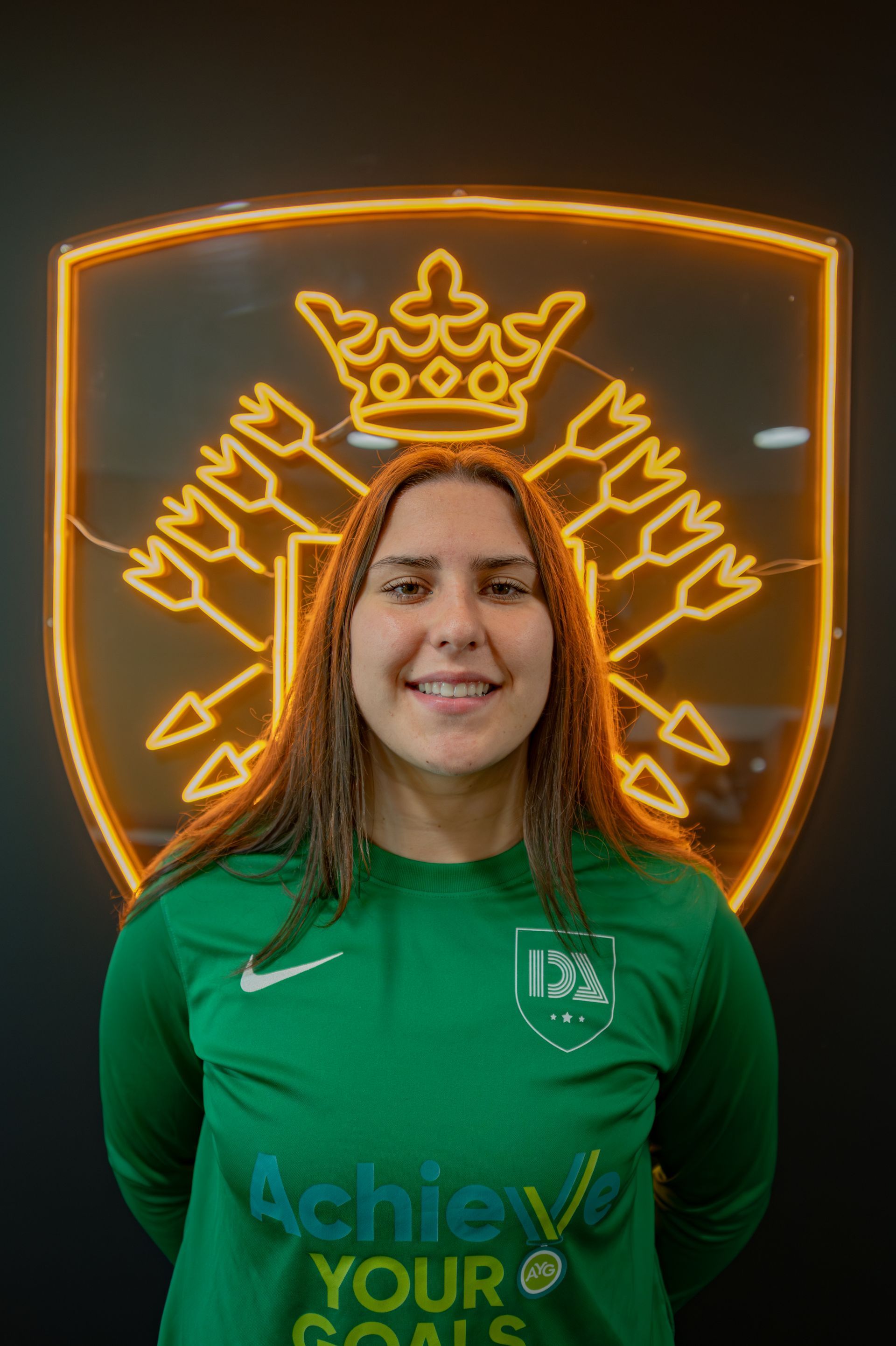 Woman in green jersey smiles, stands before a neon crest.