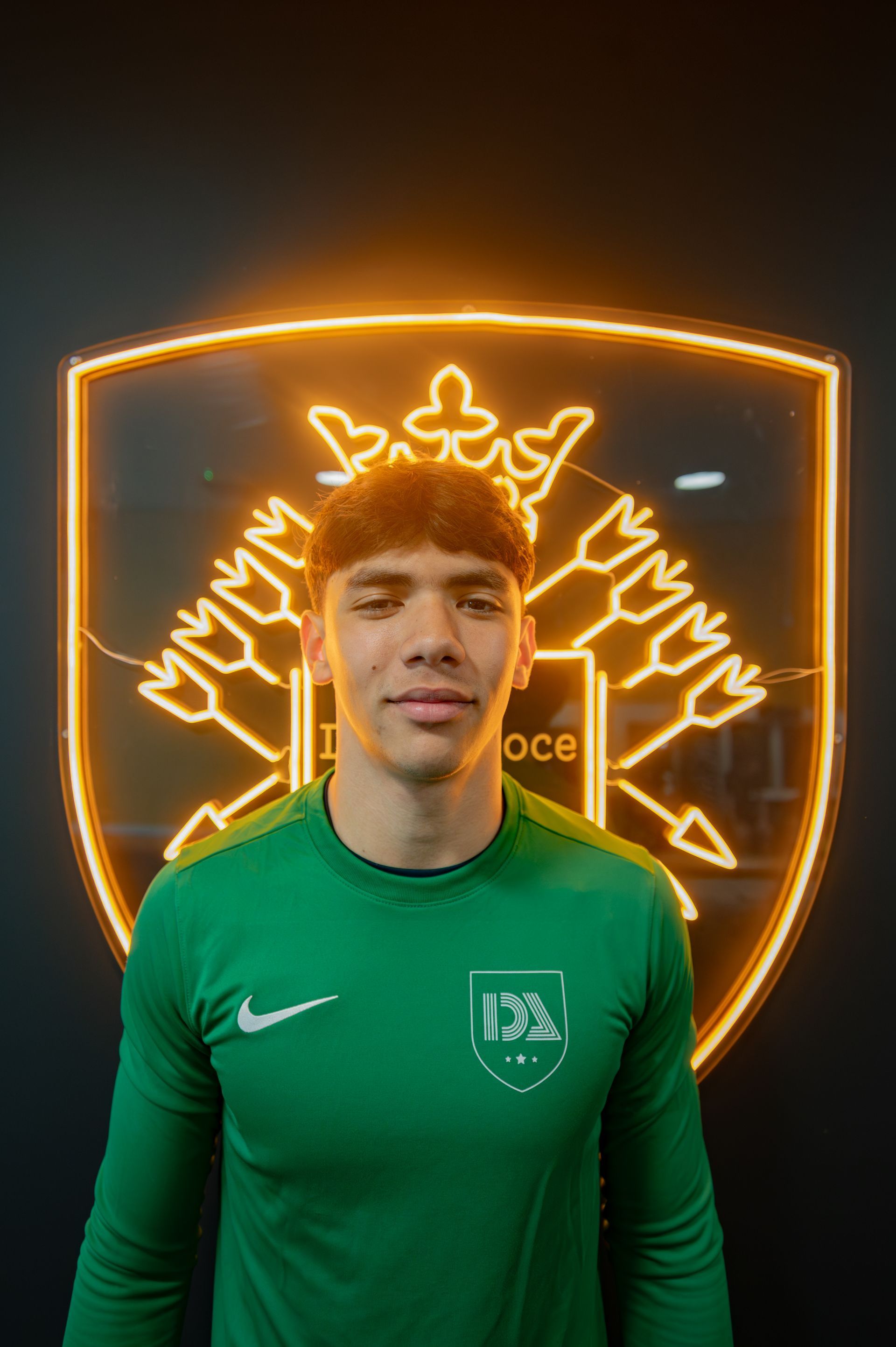 Man in a green shirt, standing in front of a team crest illuminated with an orange glow.