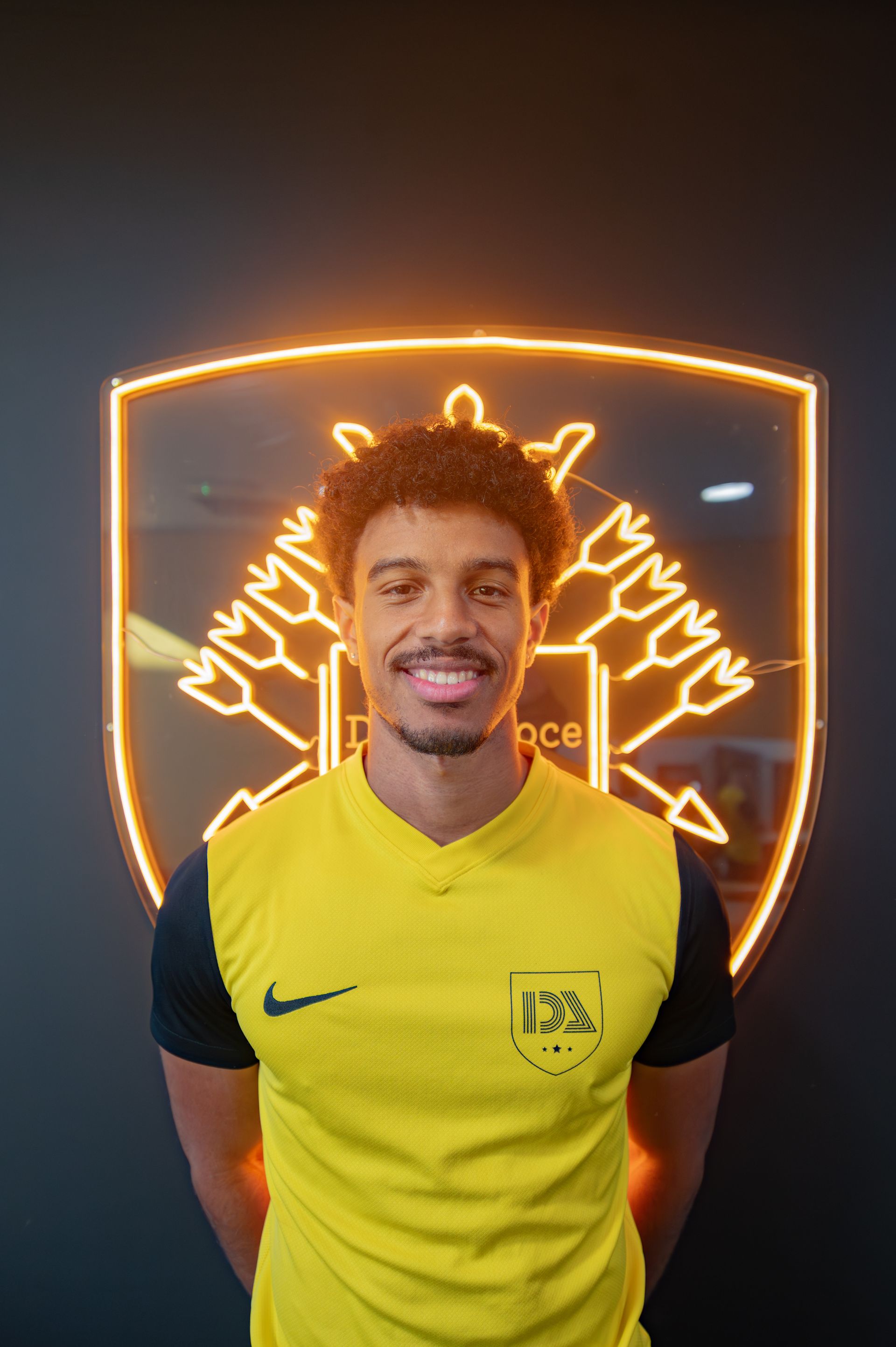 Soccer player smiling, wearing yellow jersey with logo, standing in front of a neon team crest.