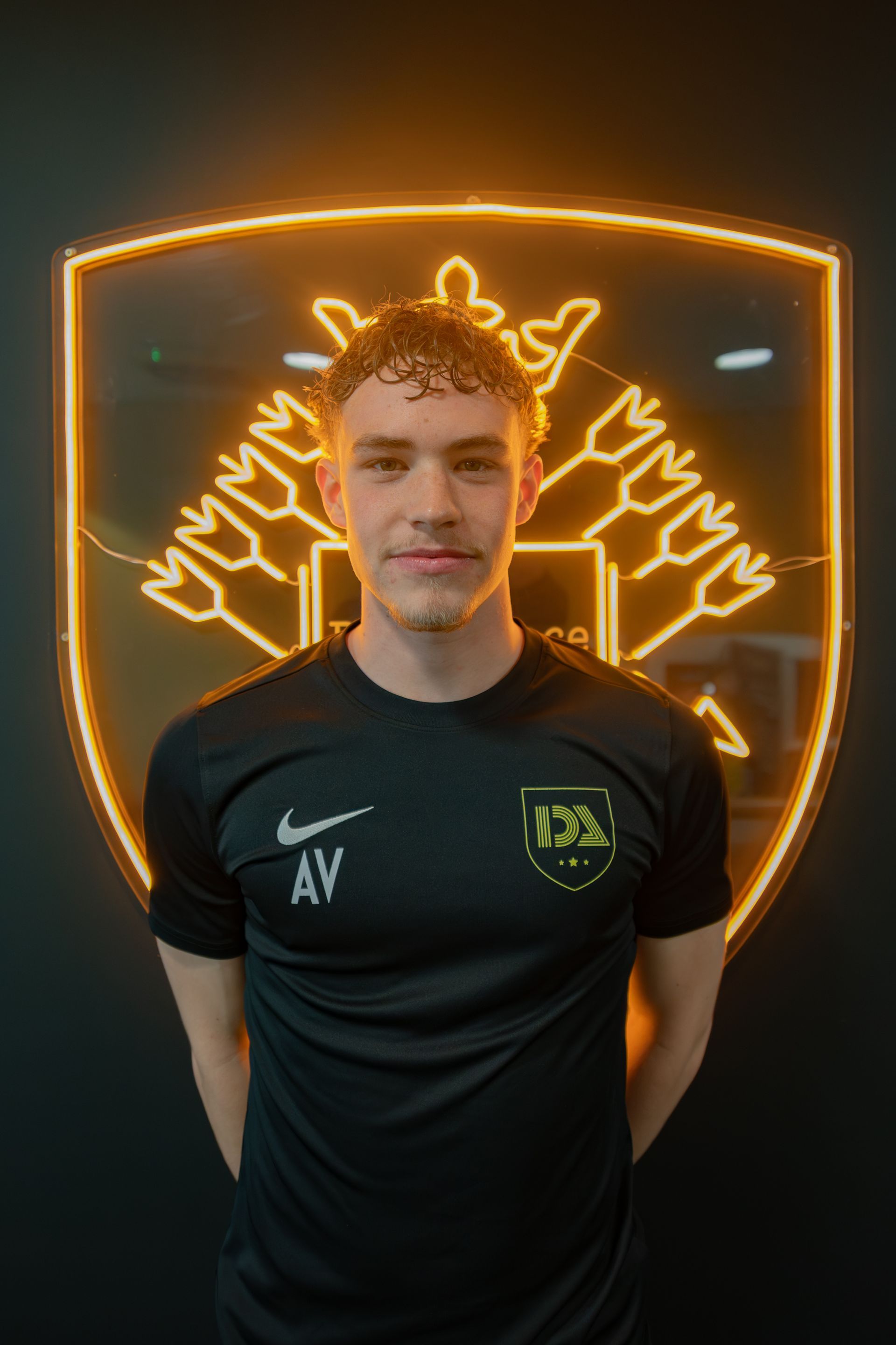 Man in black shirt with logo, stands in front of a neon emblem.