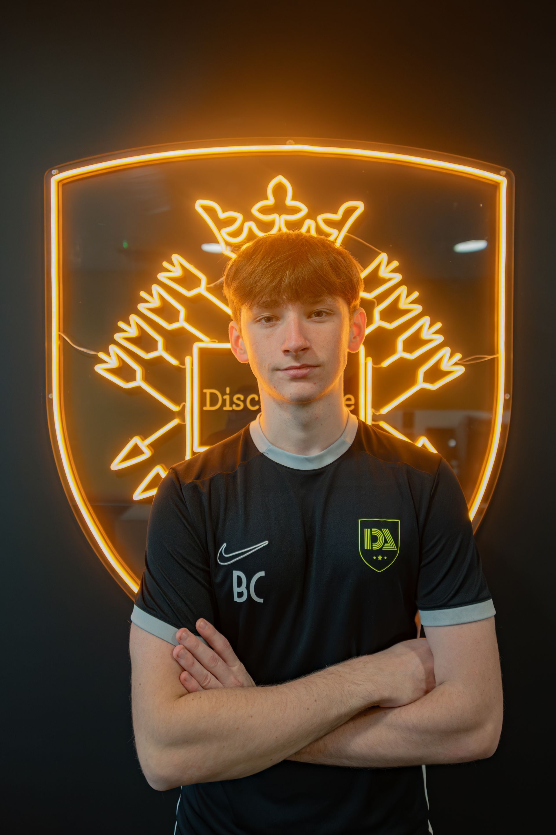Young man in black shirt with arms crossed, in front of a lit, gold-colored shield emblem.