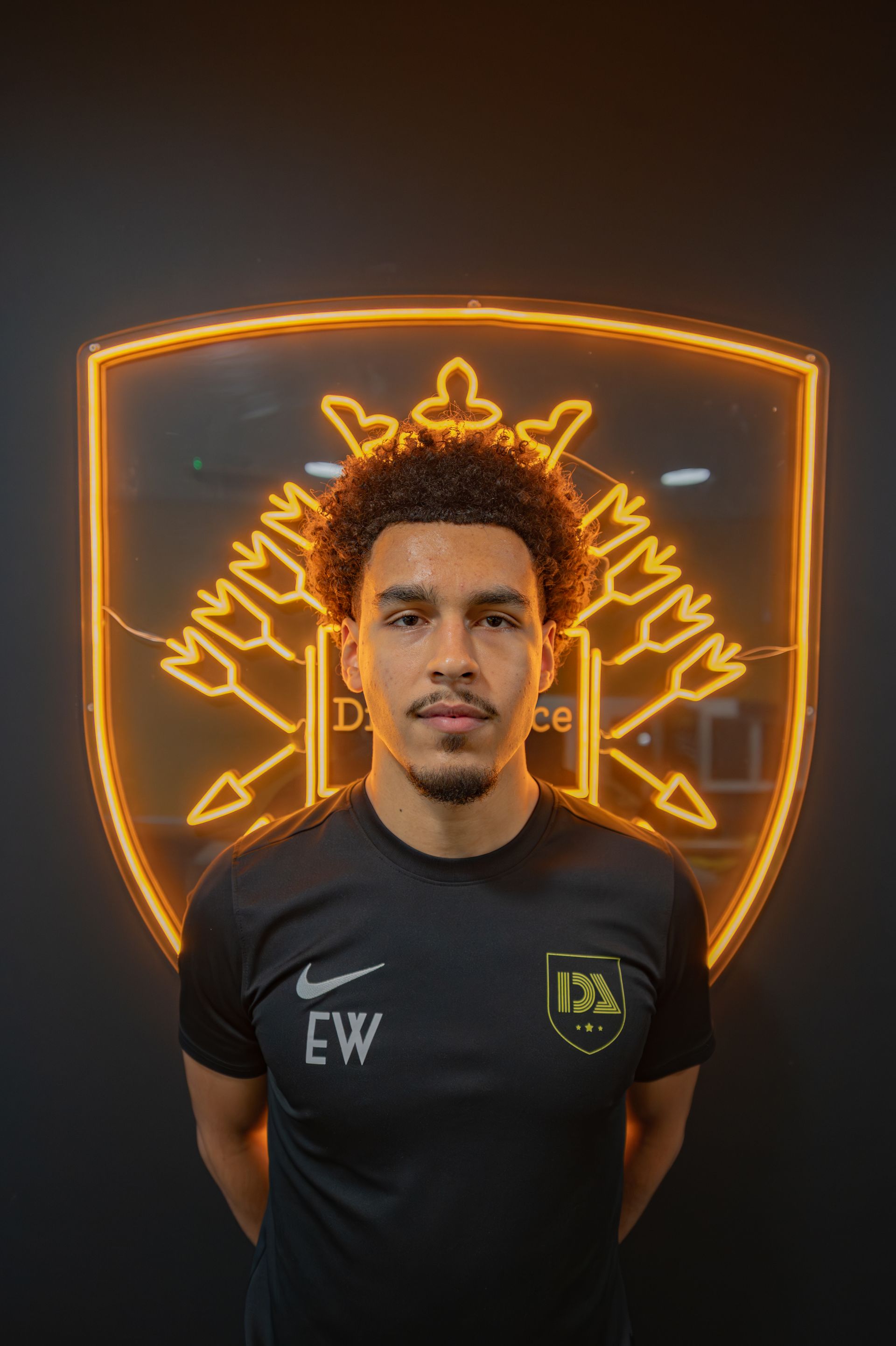 Man in black shirt stands in front of a neon logo. His curly hair and focused expression.