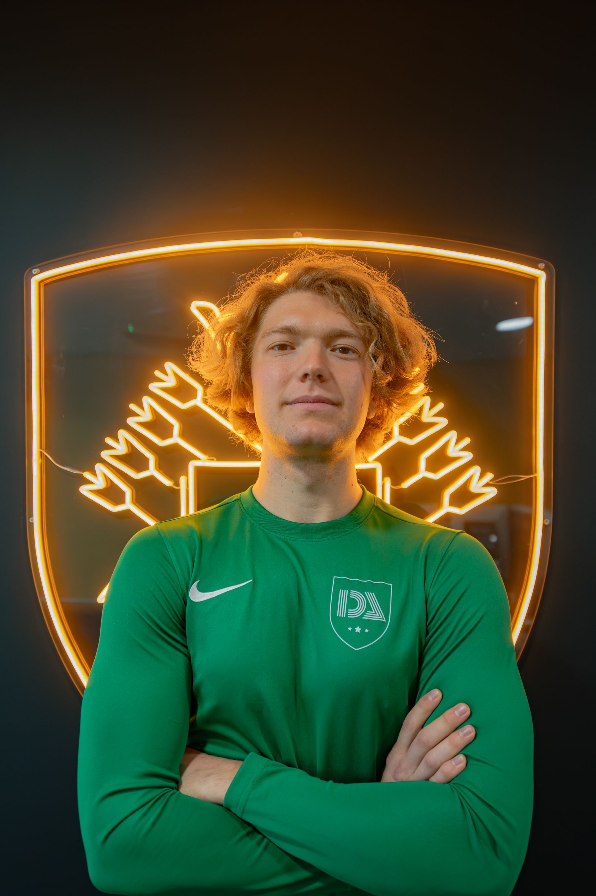Man in green shirt, arms crossed, stands before a neon logo.
