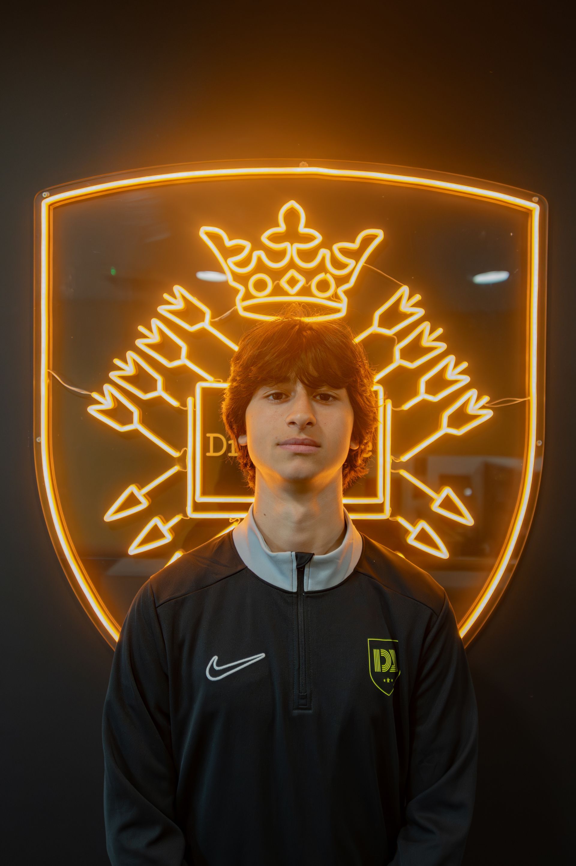 Young person stands in front of a glowing logo. They wear a black and green athletic top with a centered zipper.