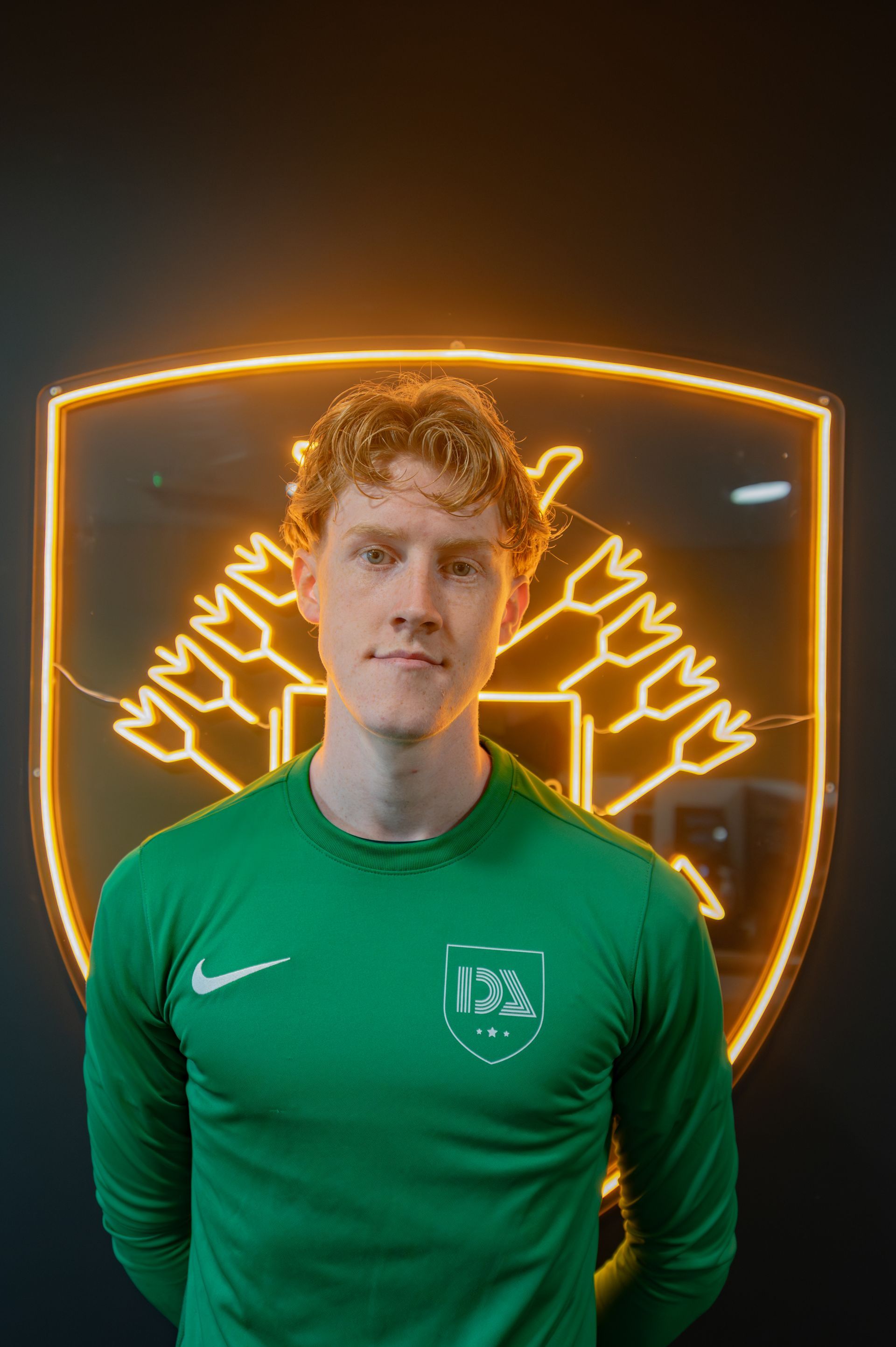 Man in green shirt, standing in front of illuminated gold emblem on black wall.