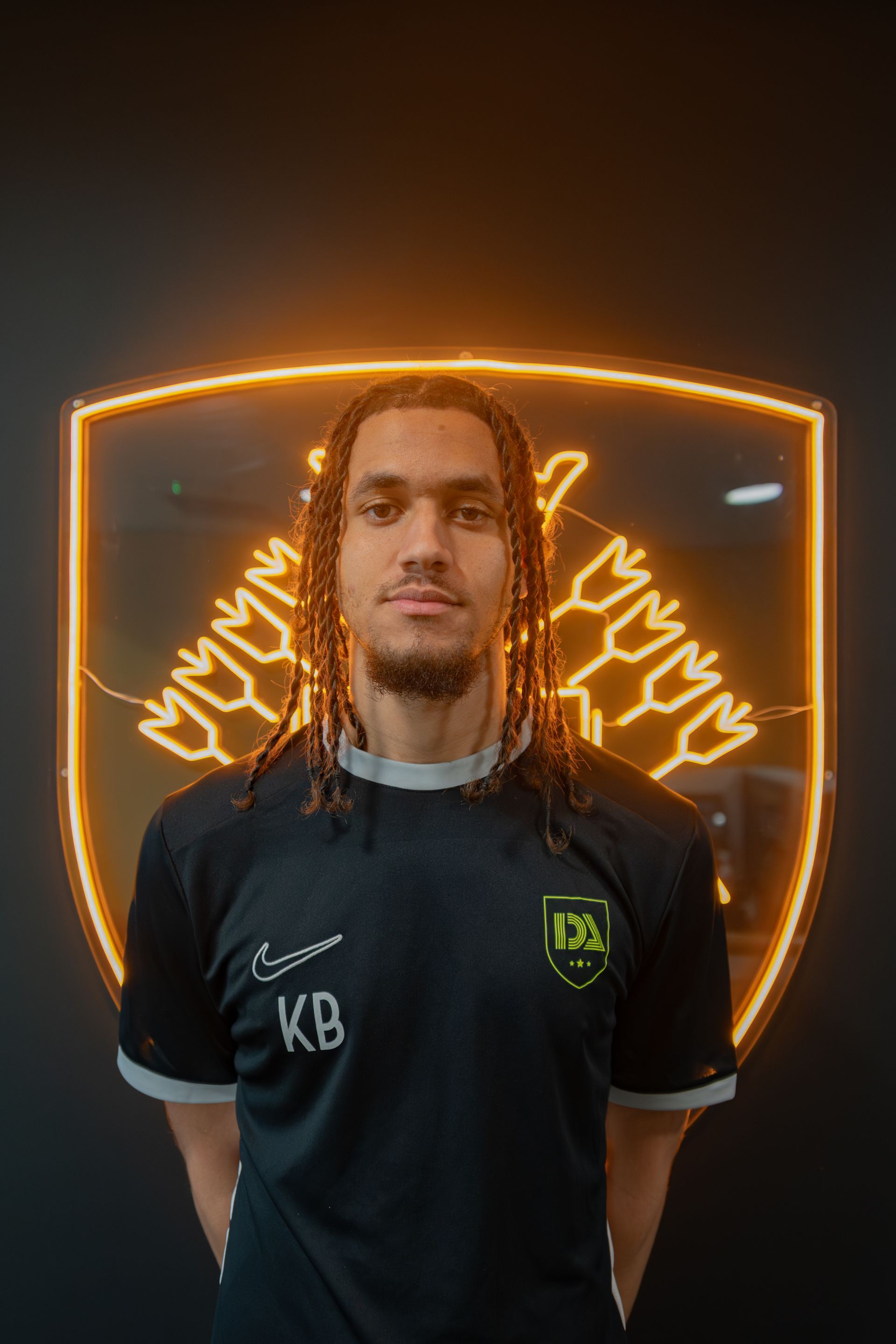 Man with dreadlocks wearing a black shirt, standing in front of a lit emblem.