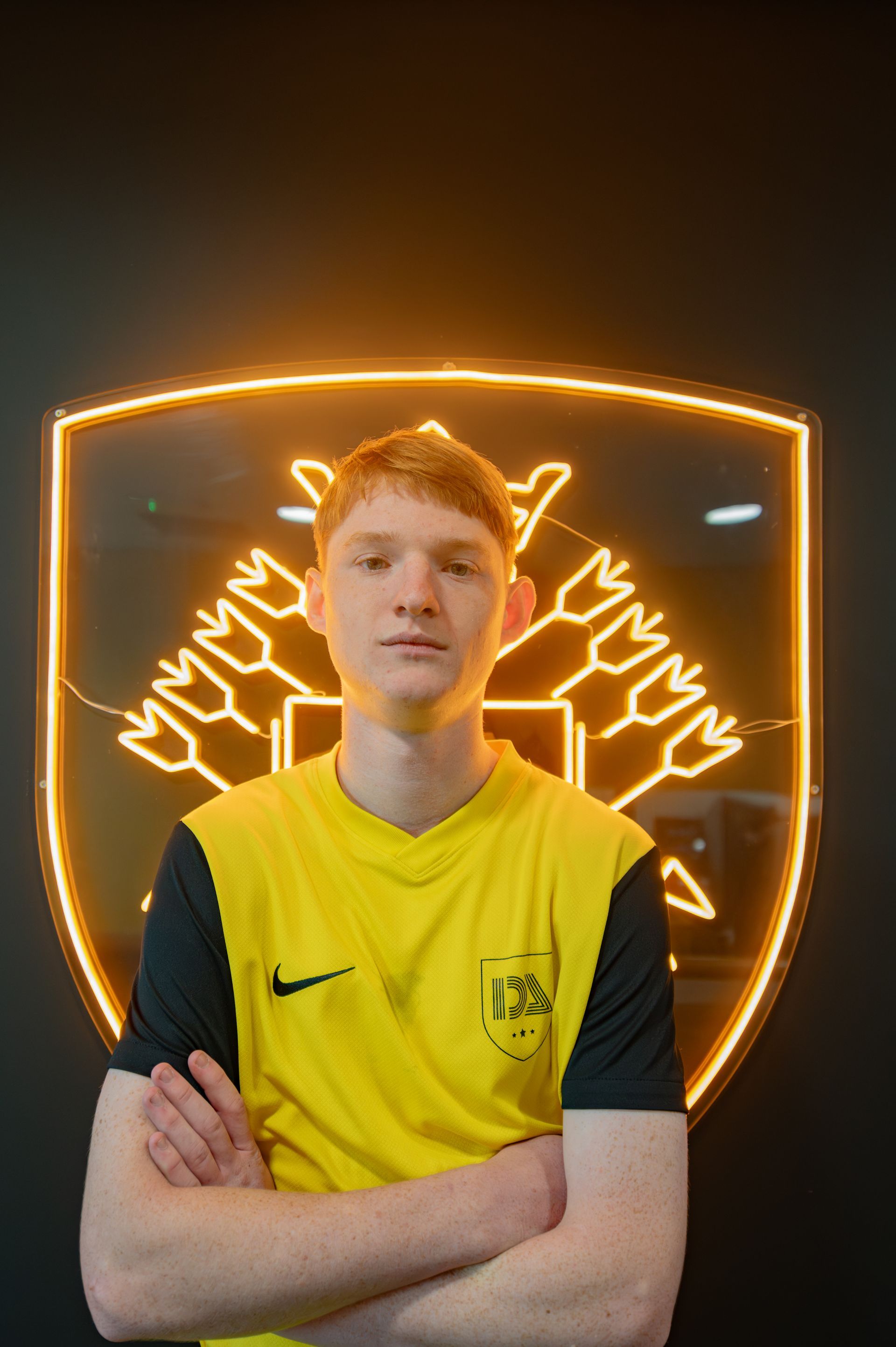 Young person in yellow and black jersey stands in front of a neon emblem. Arms are crossed.