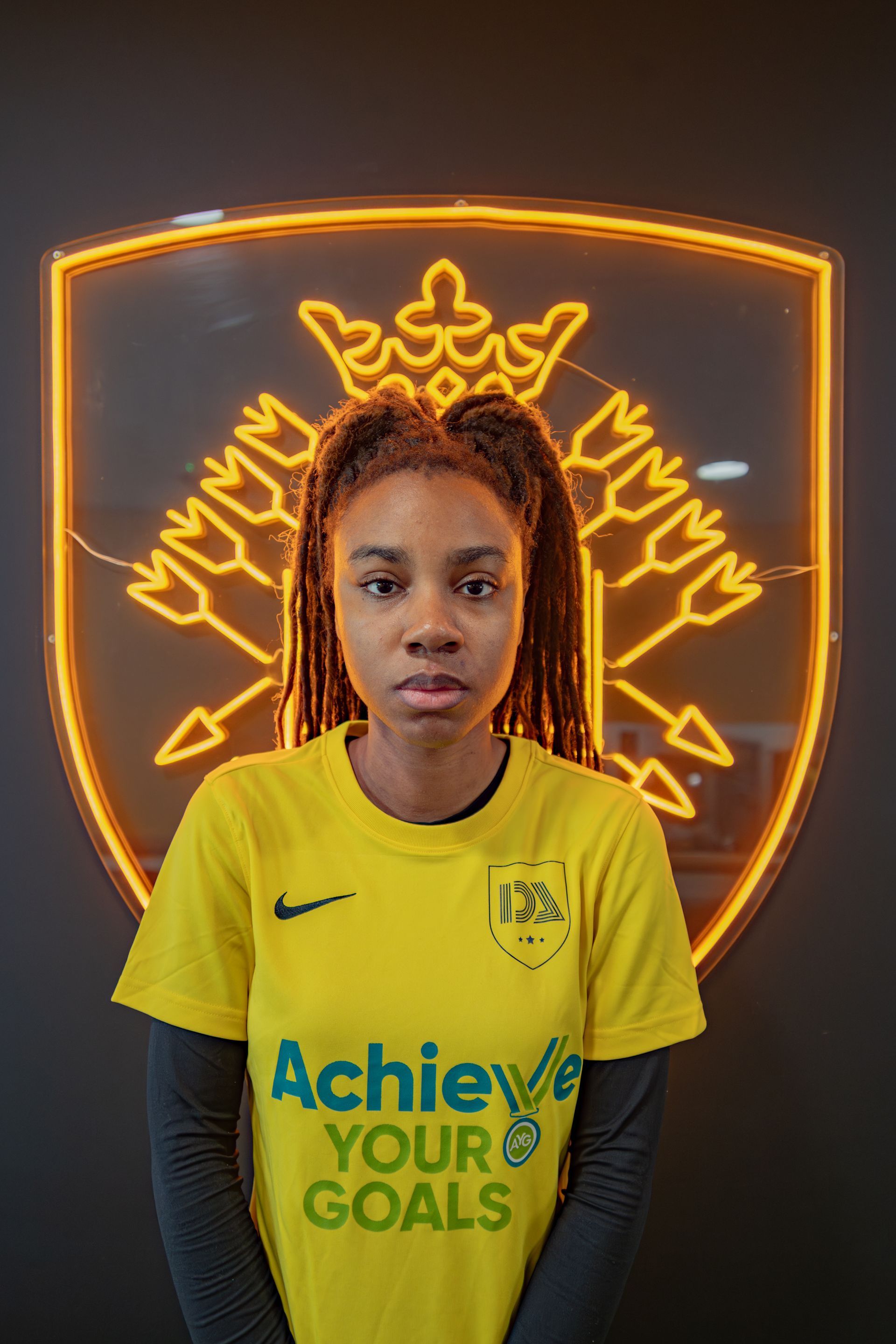Woman in yellow jersey stands before a golden logo. The jersey says 