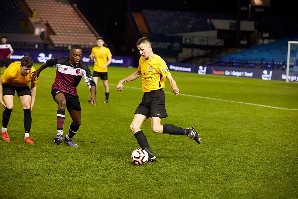 Soccer players in yellow and maroon uniforms on a green field at night, one kicking the ball.