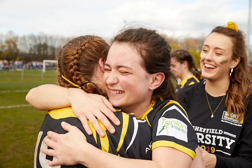Two women in sports jerseys hug, smiling, on a field. Another woman smiles nearby.