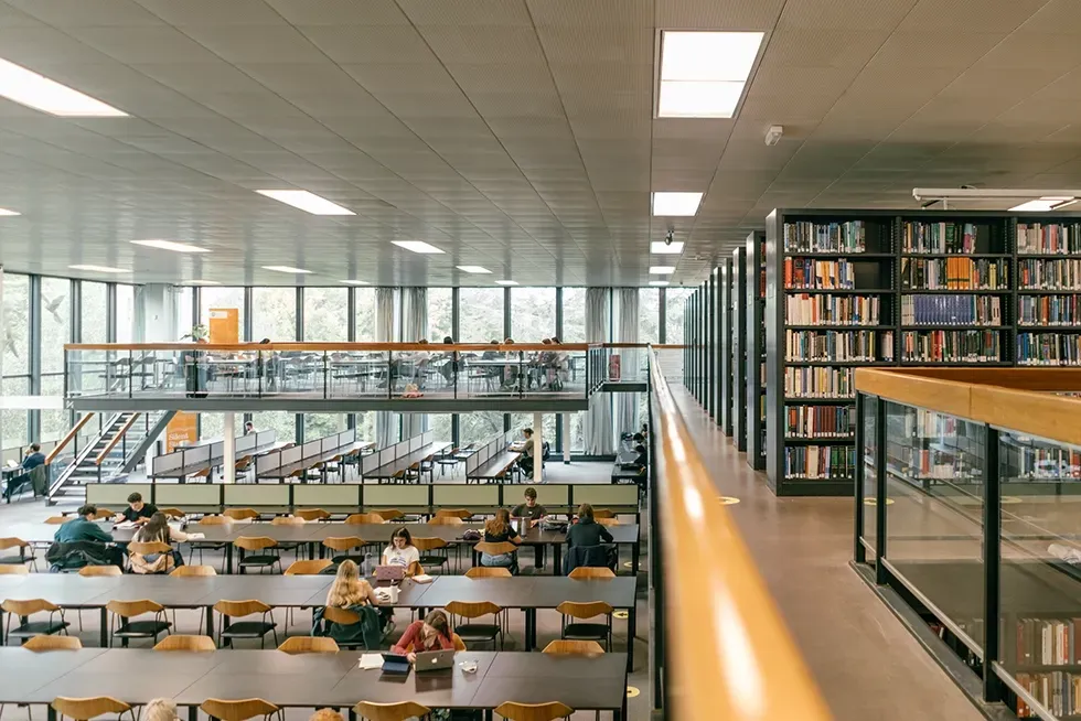 Library interior with rows of tables, bookshelves, and a mezzanine level. Natural light streams in.