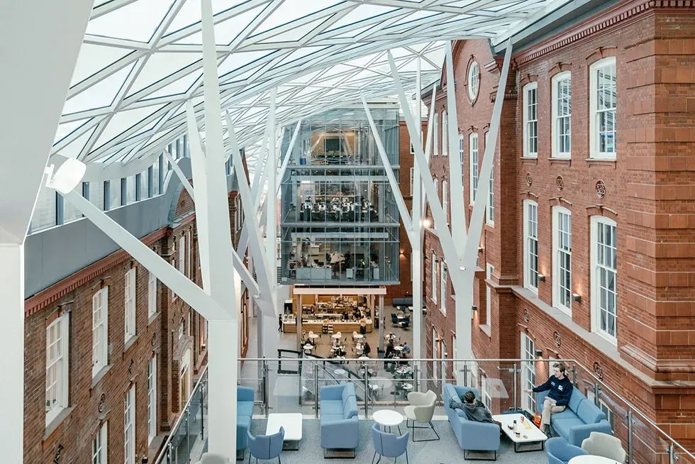 Interior view of a modern building with glass roof, brick walls, and white support beams. People sit in lounge areas.