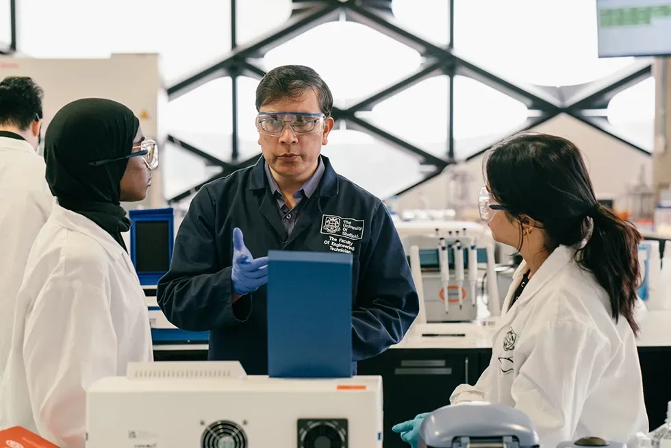 A lab instructor gestures, speaking to two students wearing lab coats and safety glasses. Bright laboratory setting.