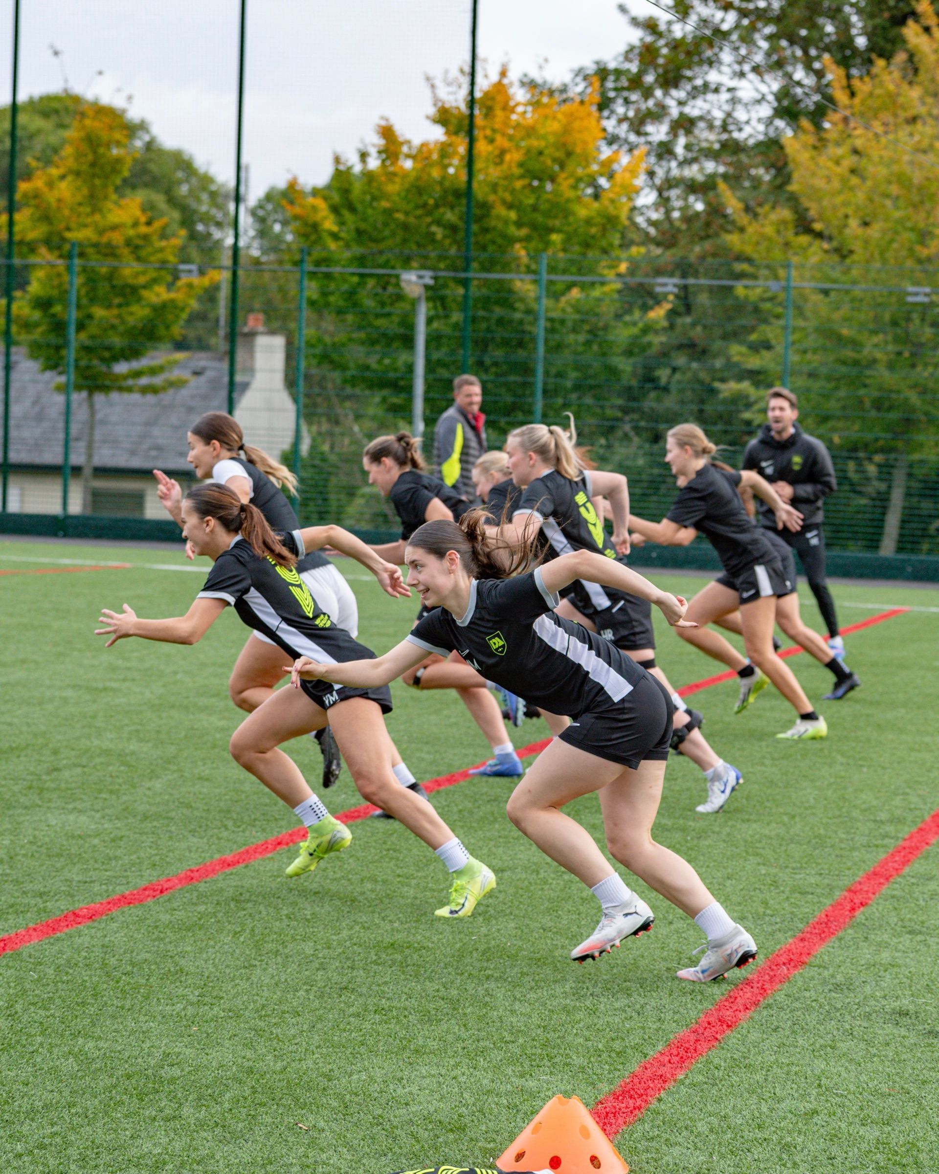 Women in black athletic gear sprint on a green field with red lines.