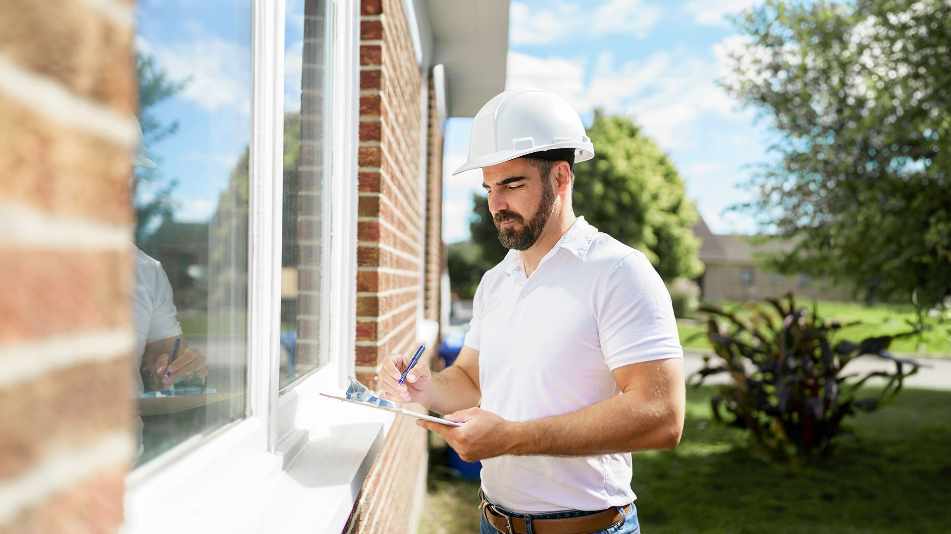 Worker in a white hard hat writing notes beside a brick building window outside.