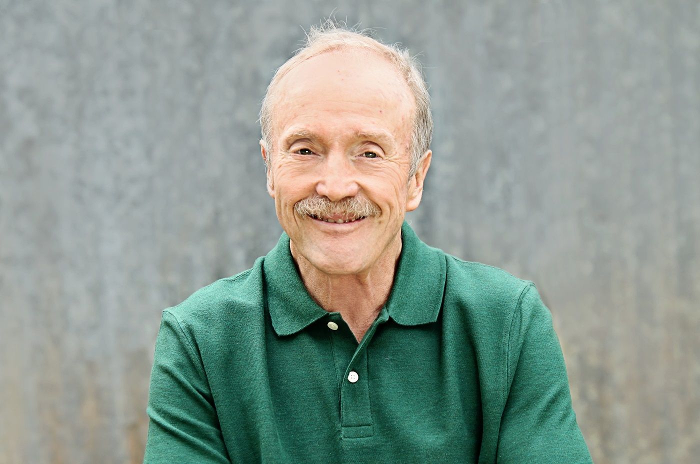 Man with mustache wearing a green polo shirt smiling, set against a gray background.