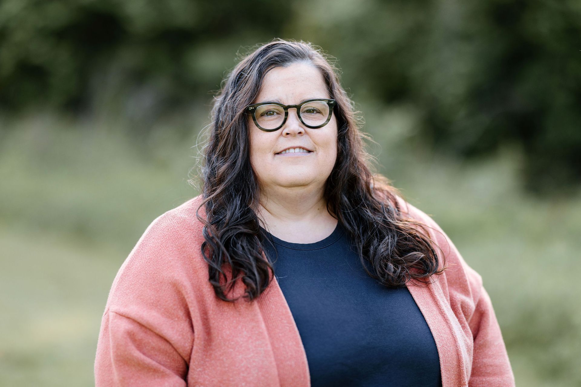 Woman wearing glasses and a coral cardigan smiles outdoors.
