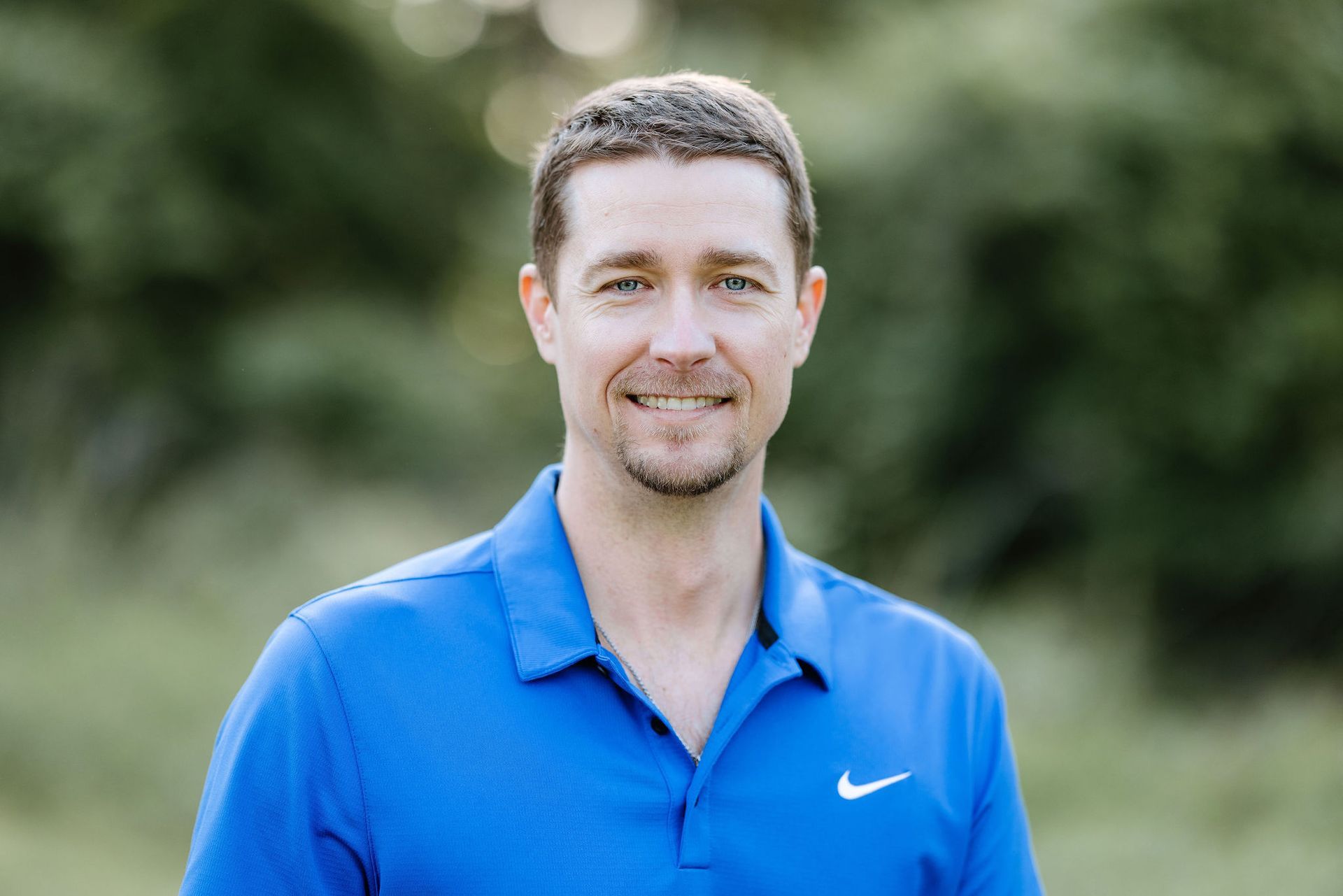 Man smiling, wearing a blue polo shirt with a Nike logo, outdoors with a green background.