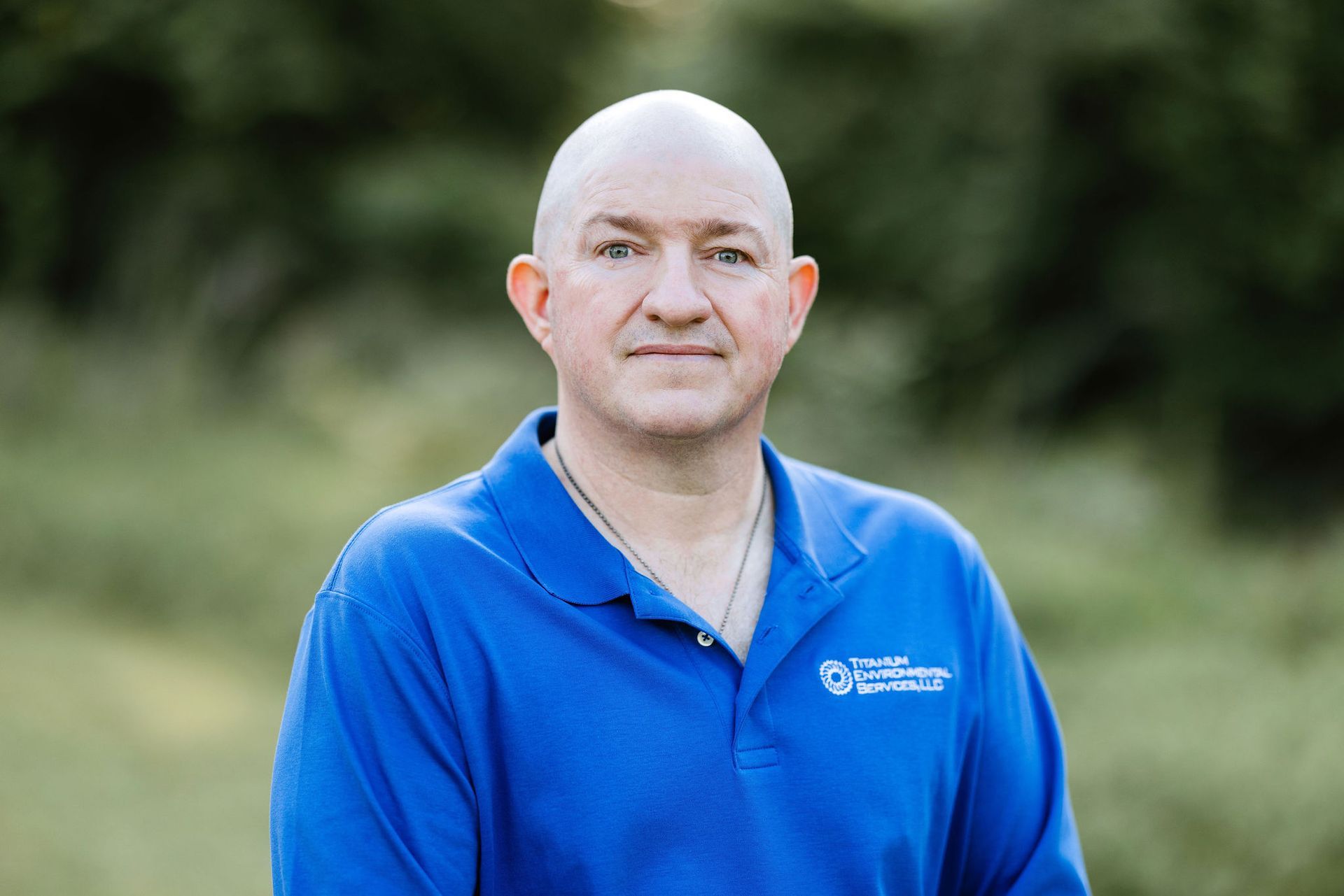Man with shaved head in a blue polo shirt, smiling slightly, outdoors with blurred green background.