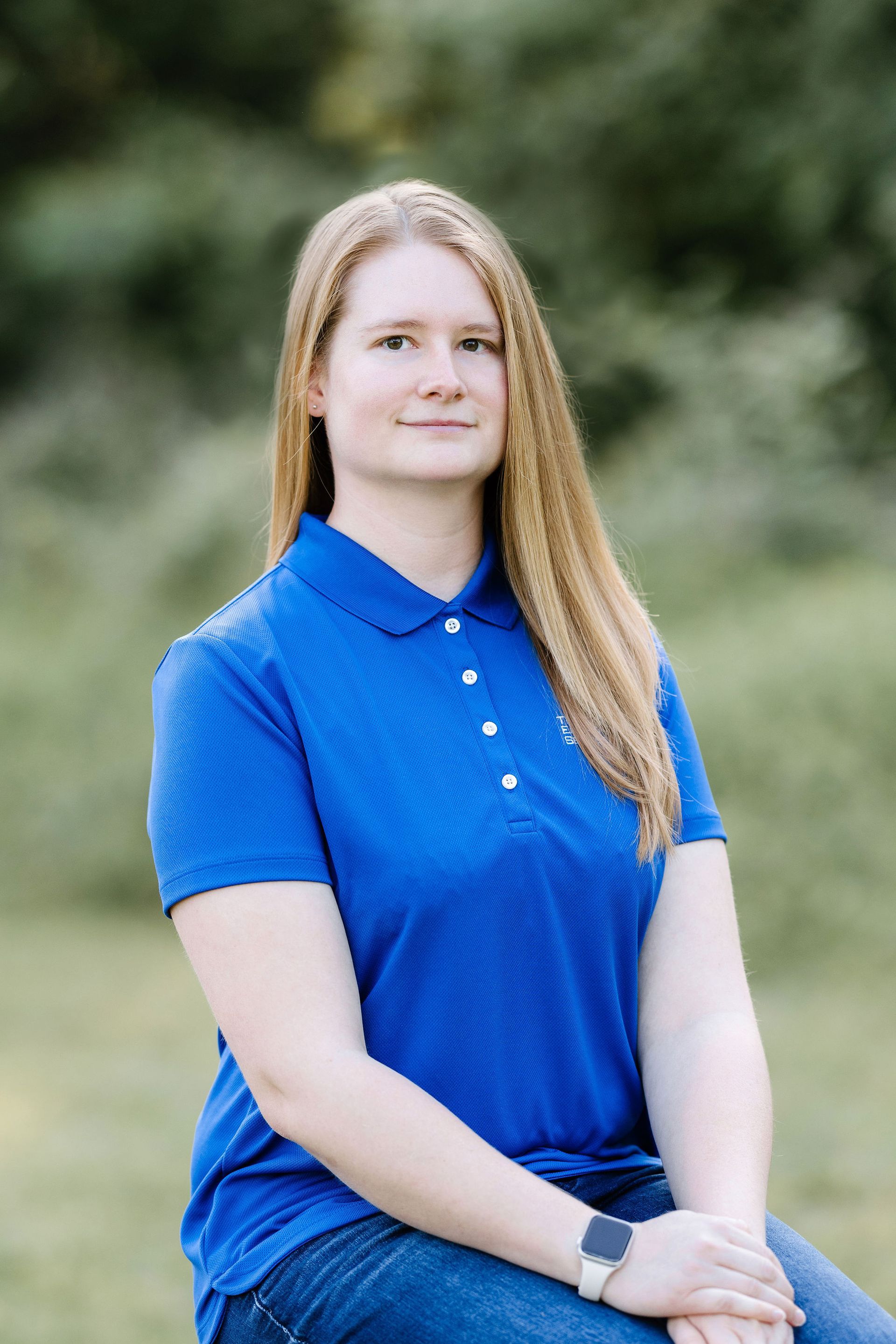 Woman with long blonde hair, blue shirt, sitting, smiling, outdoors.